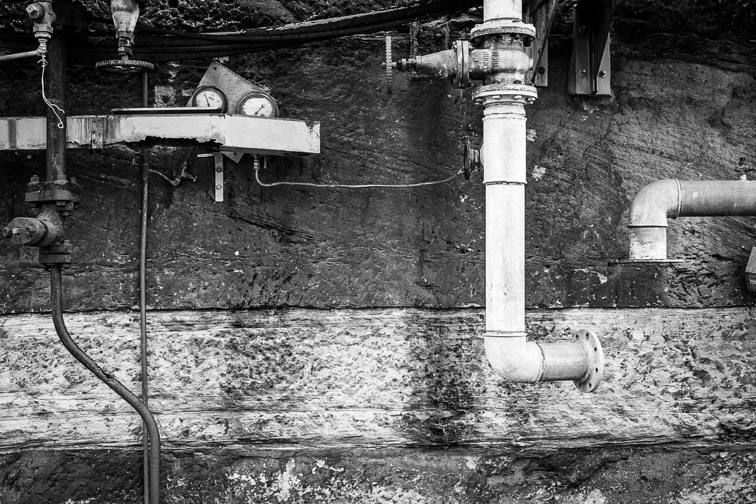 Black and white photograph of industrial pipes and gauges attached to a rock wall, with various valves and meters.