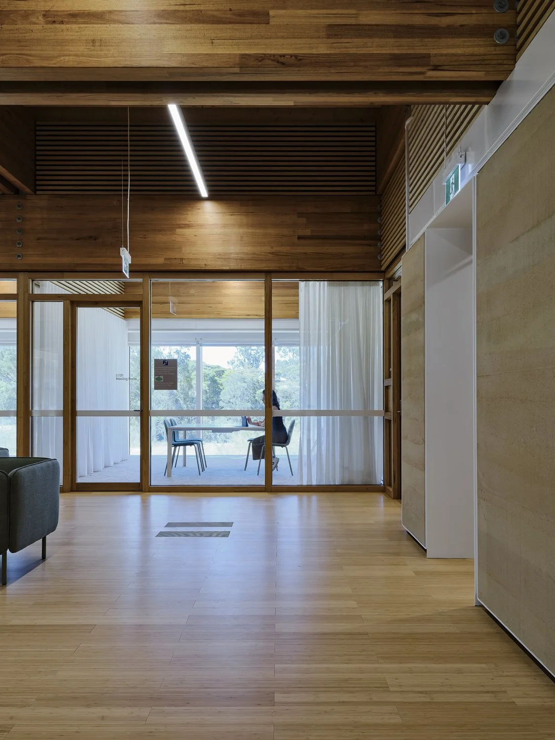 An interior view of a modern office or waiting area with wooden flooring and walls, glass partition, and white curtains; a woman is sitting at a table in a sunlit room.
