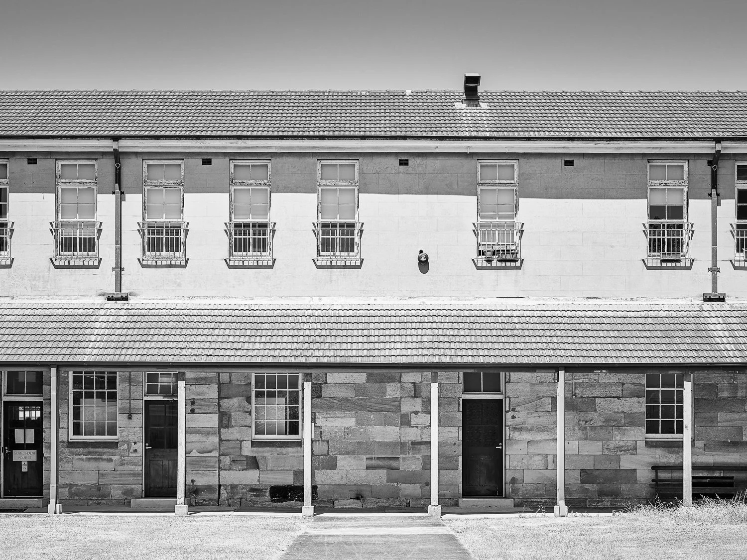A black and white photo of a two-story building with a tiled roof. The upper story has six windows with small balconies, and the lower story has three doors and three windows, with vertical posts supporting a porch roof.