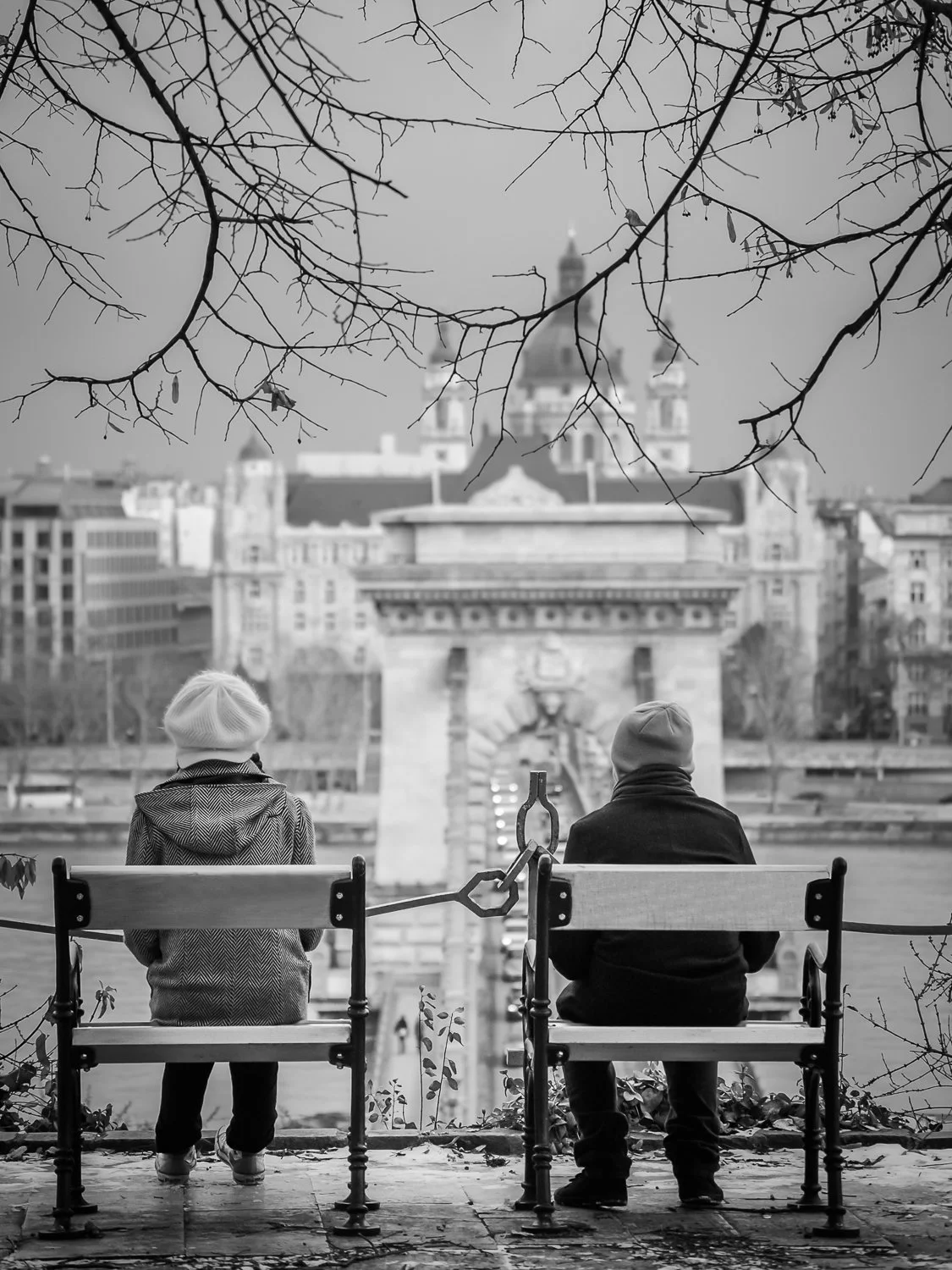 Two people sitting on benches, facing the Pont Alexandre III bridge in Paris, France, with leafless tree branches overhead and a cityscape in the background, in black and white.