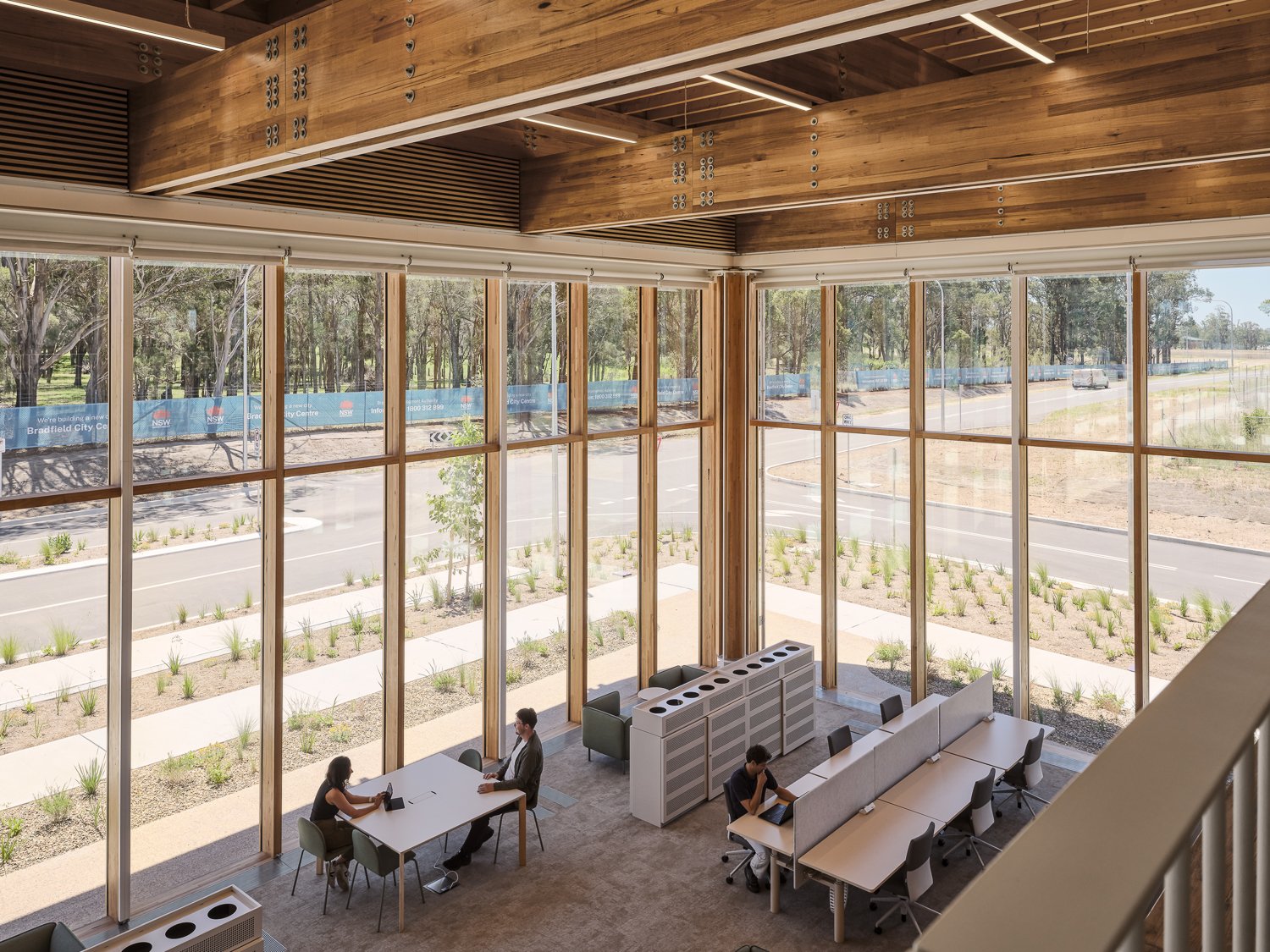 Interior of a modern office space with tall windows, wooden beams, and several people working at desks. Outside, there is a view of a road and trees.