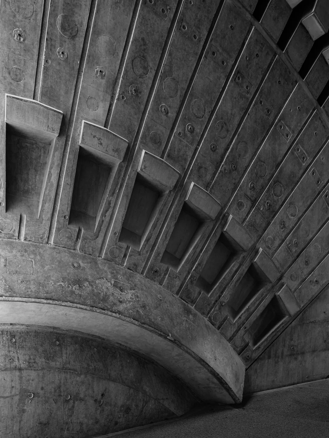 A black and white photograph of an industrial concrete structure, showing curved concrete steps and a ceiling with rectangular recesses and circular marks.