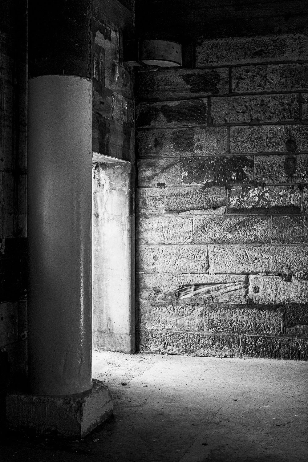 Black and white photo of a rough brick wall and concrete floor, with a tall cylindrical column on the left side near a doorway.