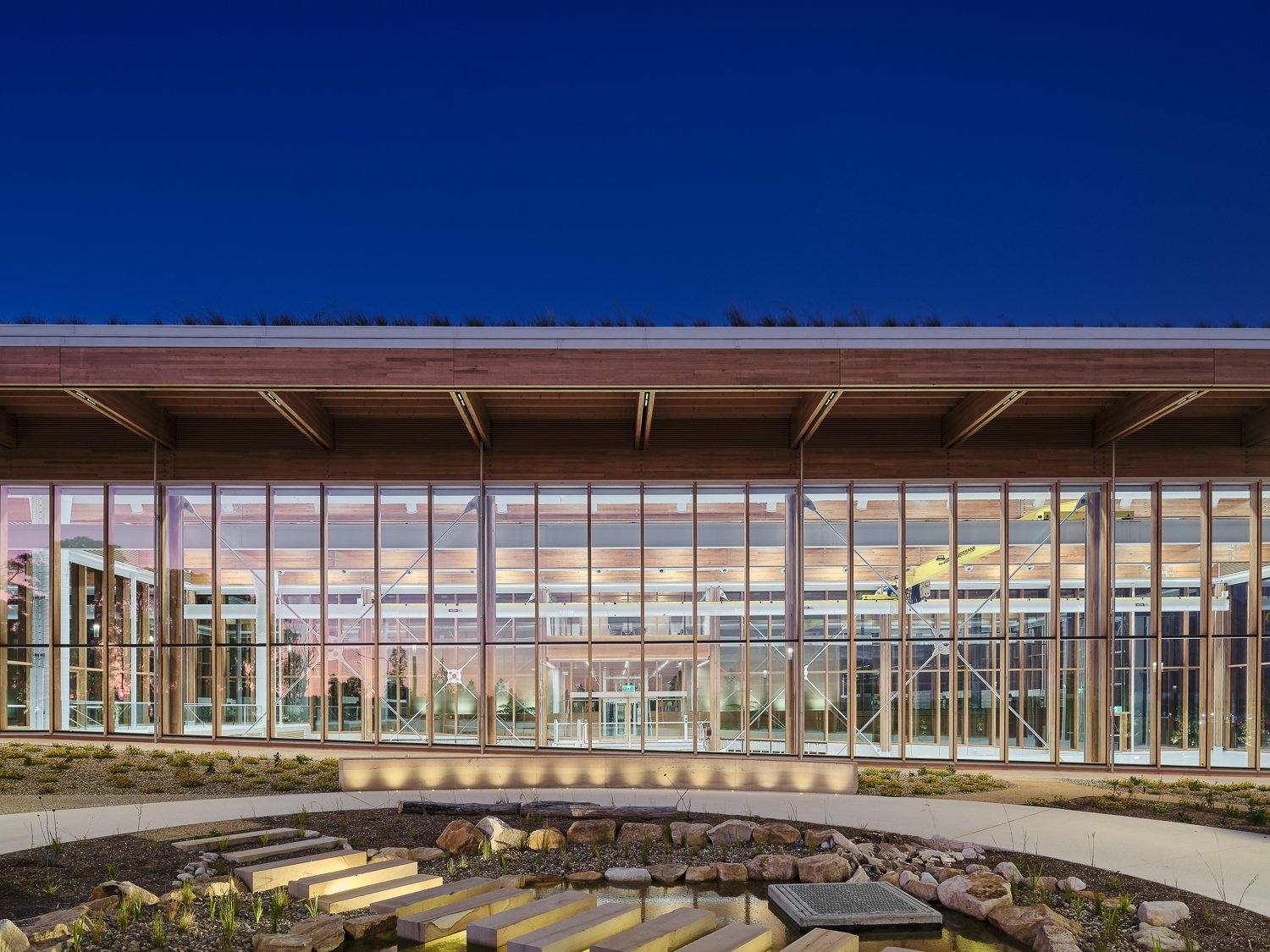Modern building with large glass windows and a wooden beam structure, surrounded by a landscaped garden with stones and a pathway.