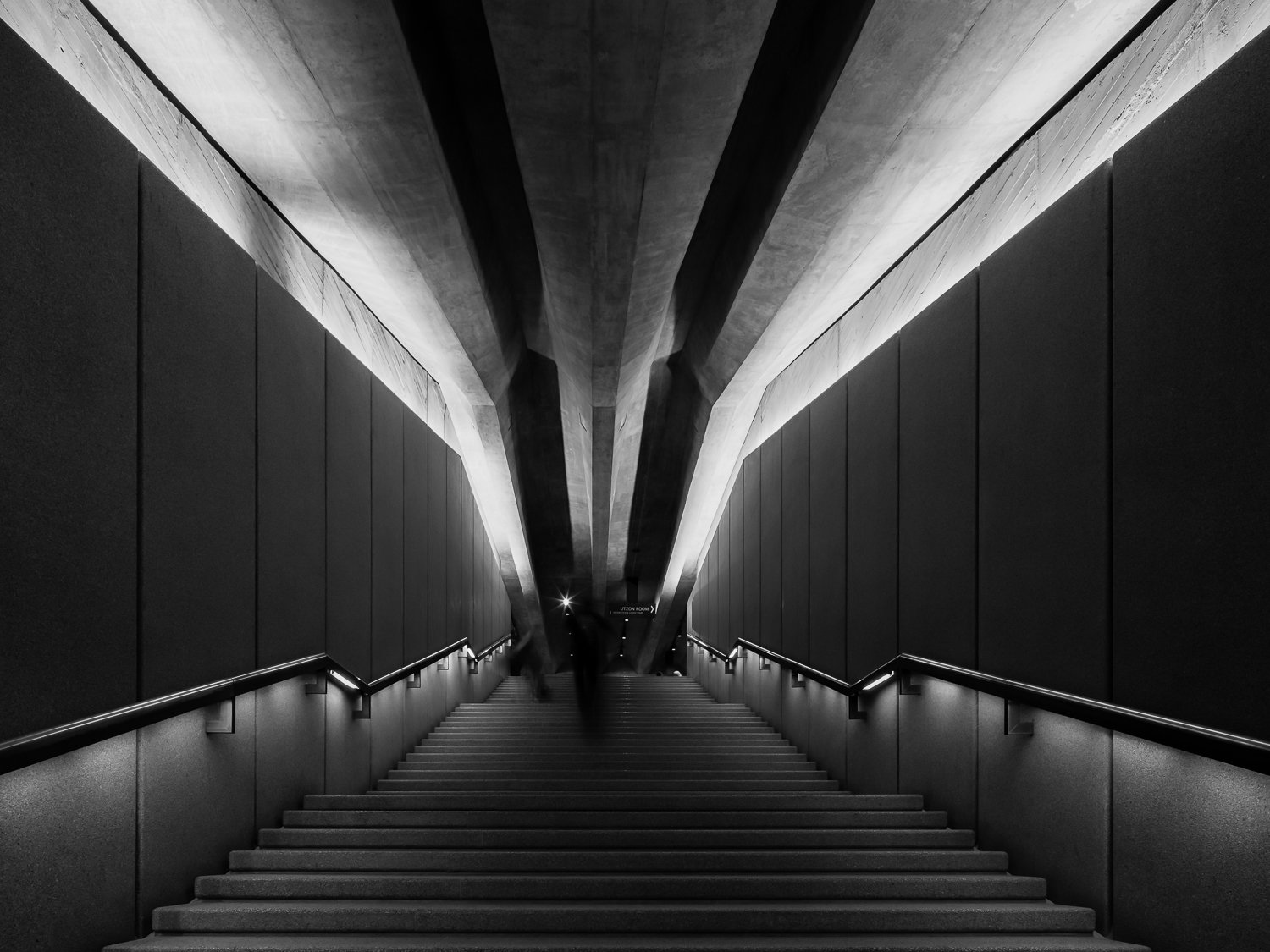 Black and white photograph of a modern staircase leading downward with sleek railings on both sides, dramatic lighting highlighting the angular architecture overhead, and a person blurred walking in the distance.