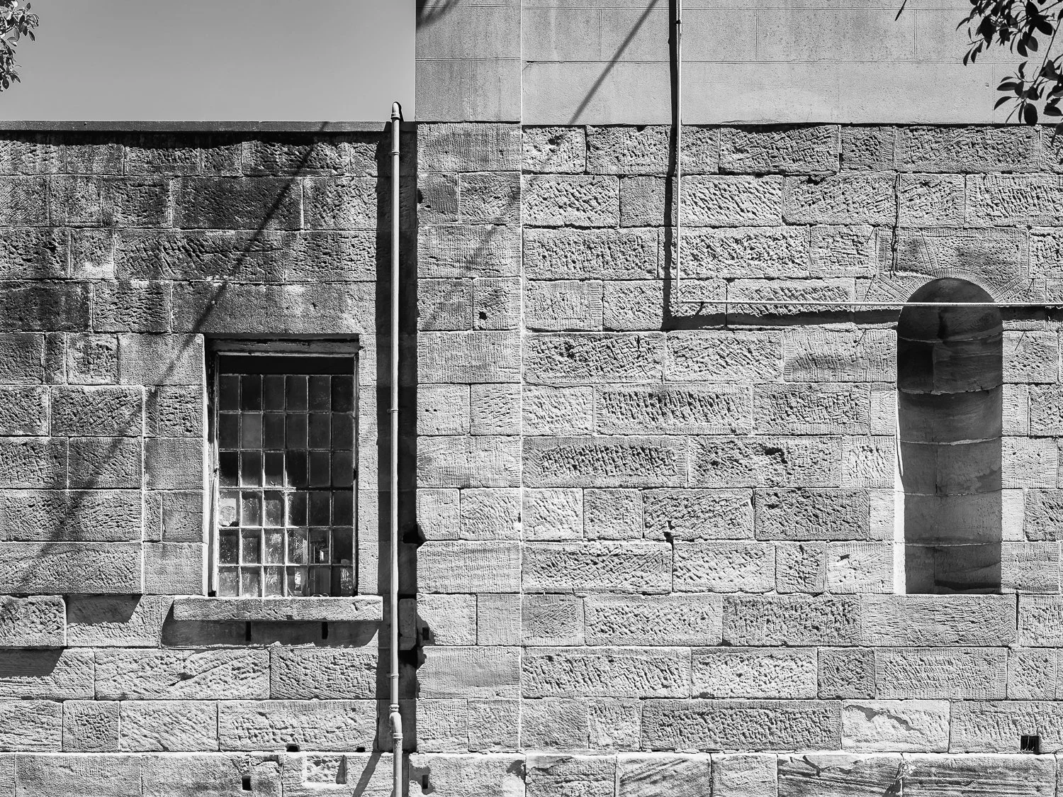 The image shows a black-and-white photo of a stone wall with two windows, one with glass blocks and the other an arched opening, and a vertical pipe running down the middle.