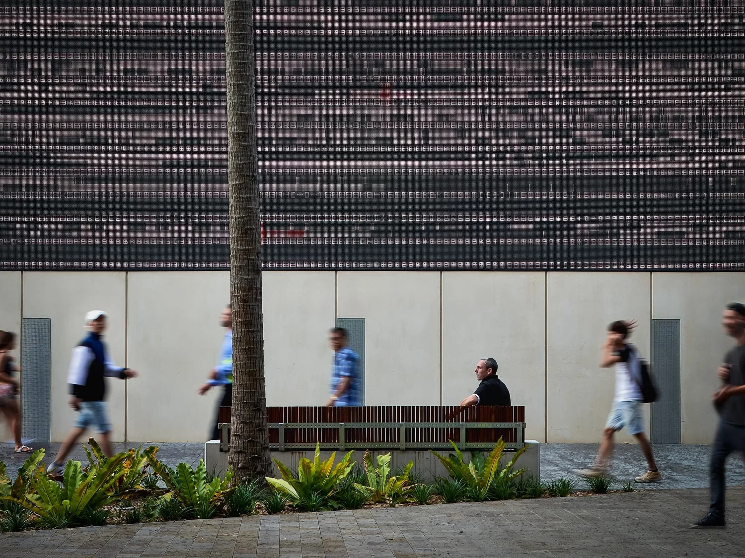 People walking on a sidewalk in front of a building with a digital advertisement display, with a tree and plants in the foreground