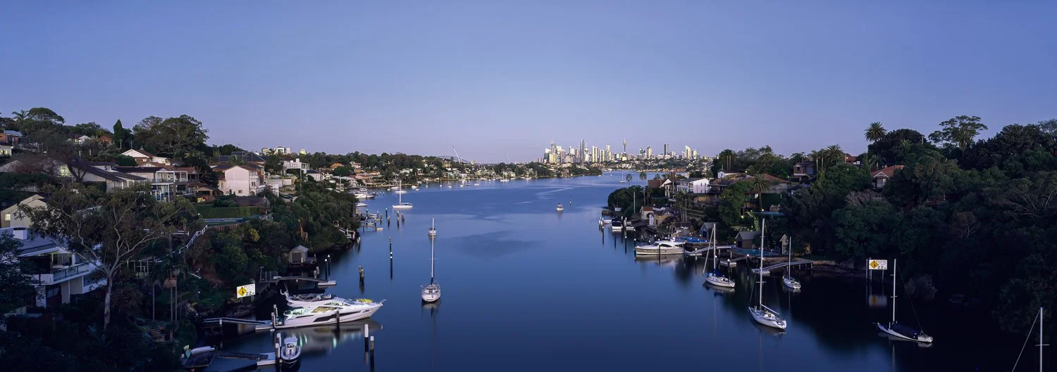 A scenic view of a wide river with boats docked along the sides, surrounded by residential houses and lush greenery, with a city skyline in the distance under a clear blue sky.