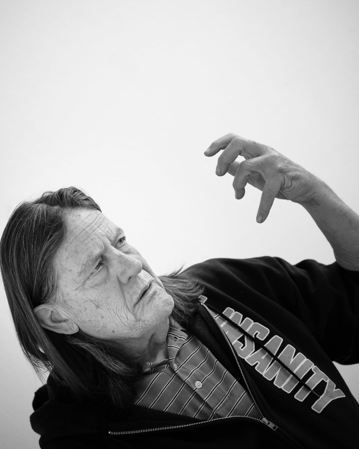 Older man with long hair and wrinkled skin looking at his hand, wearing a striped shirt and a jacket with text partially visible on it, in front of a plain background.