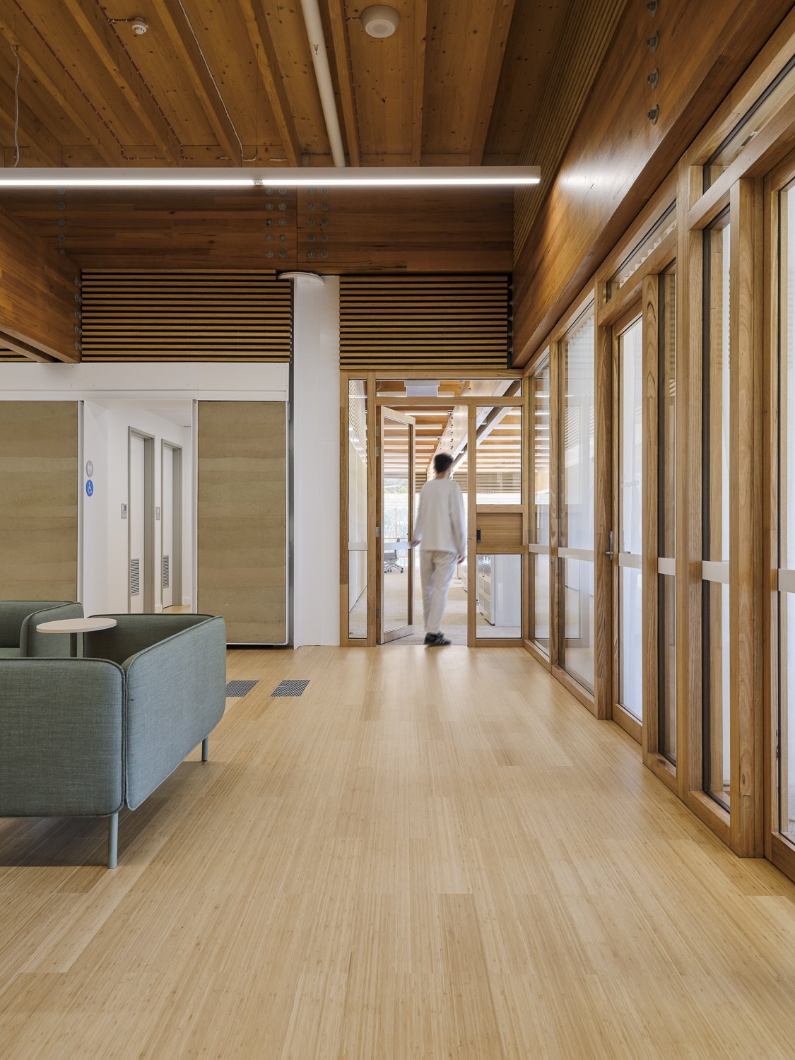 Modern interior with wooden flooring, wooden ceiling with beams, a person walking in the background, glass doors, and a green sofa in the foreground.