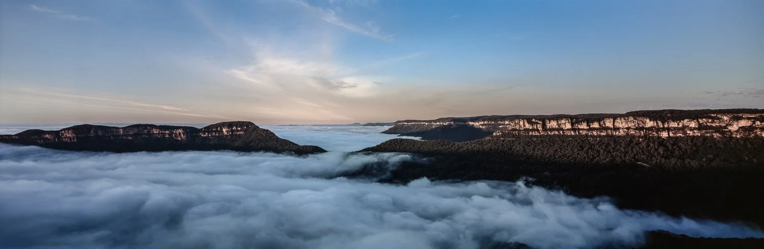 Aerial view of mountains and dense forest above a layer of clouds during sunrise or sunset.