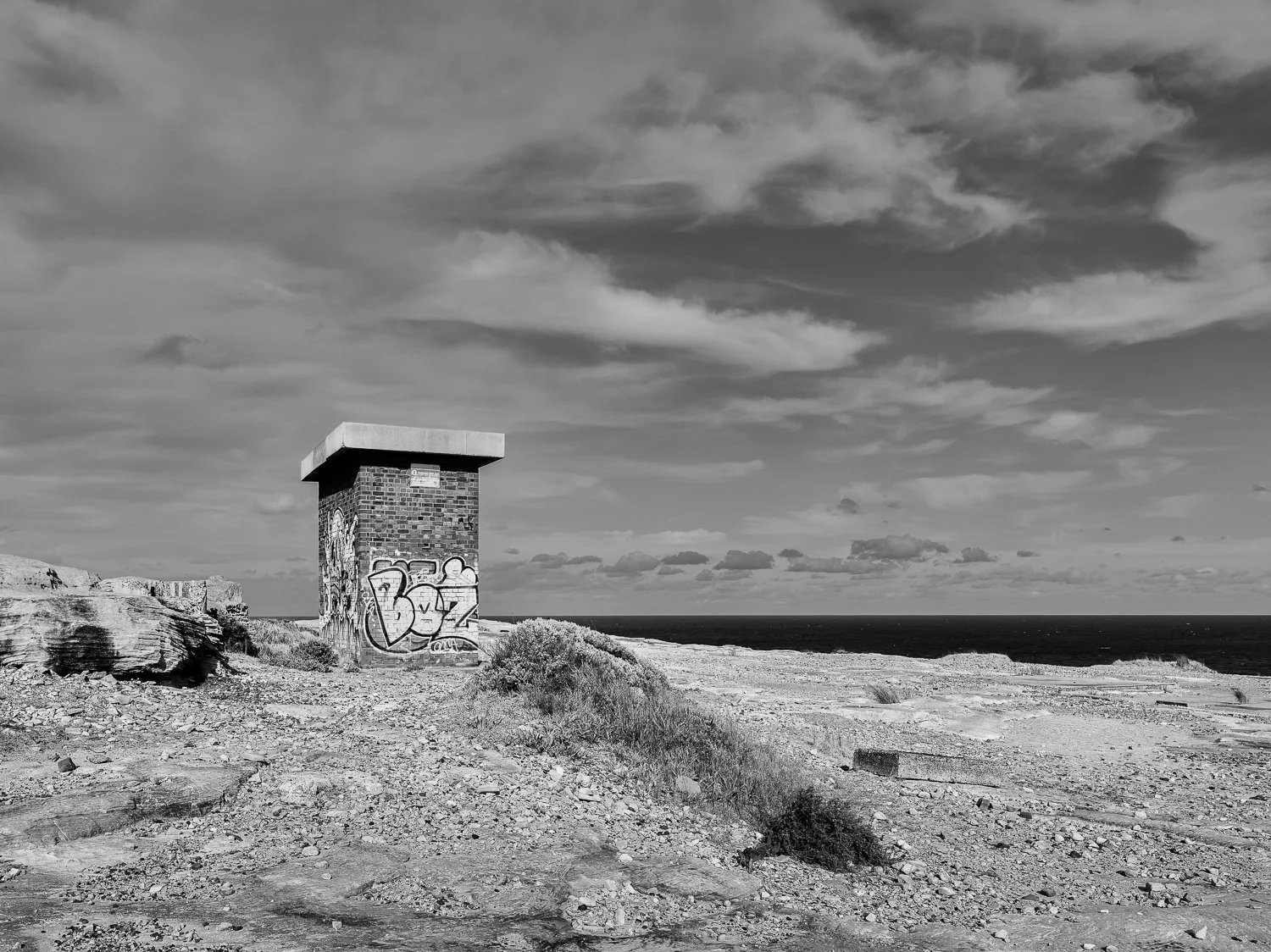 A black and white photo of a small brick structure with graffiti, situated on a rocky coastal landscape with the ocean and a cloudy sky in the background.