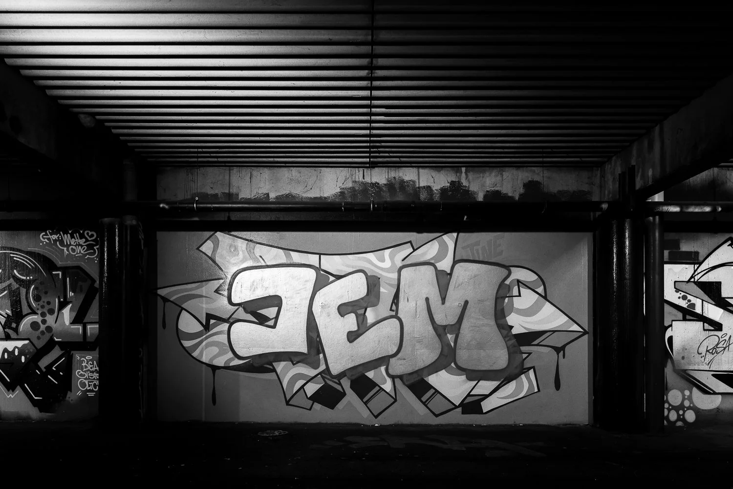 Black and white photo of graffiti on a wall under a bridge or tunnel, with the word "JEM" in bold, stylized letters.