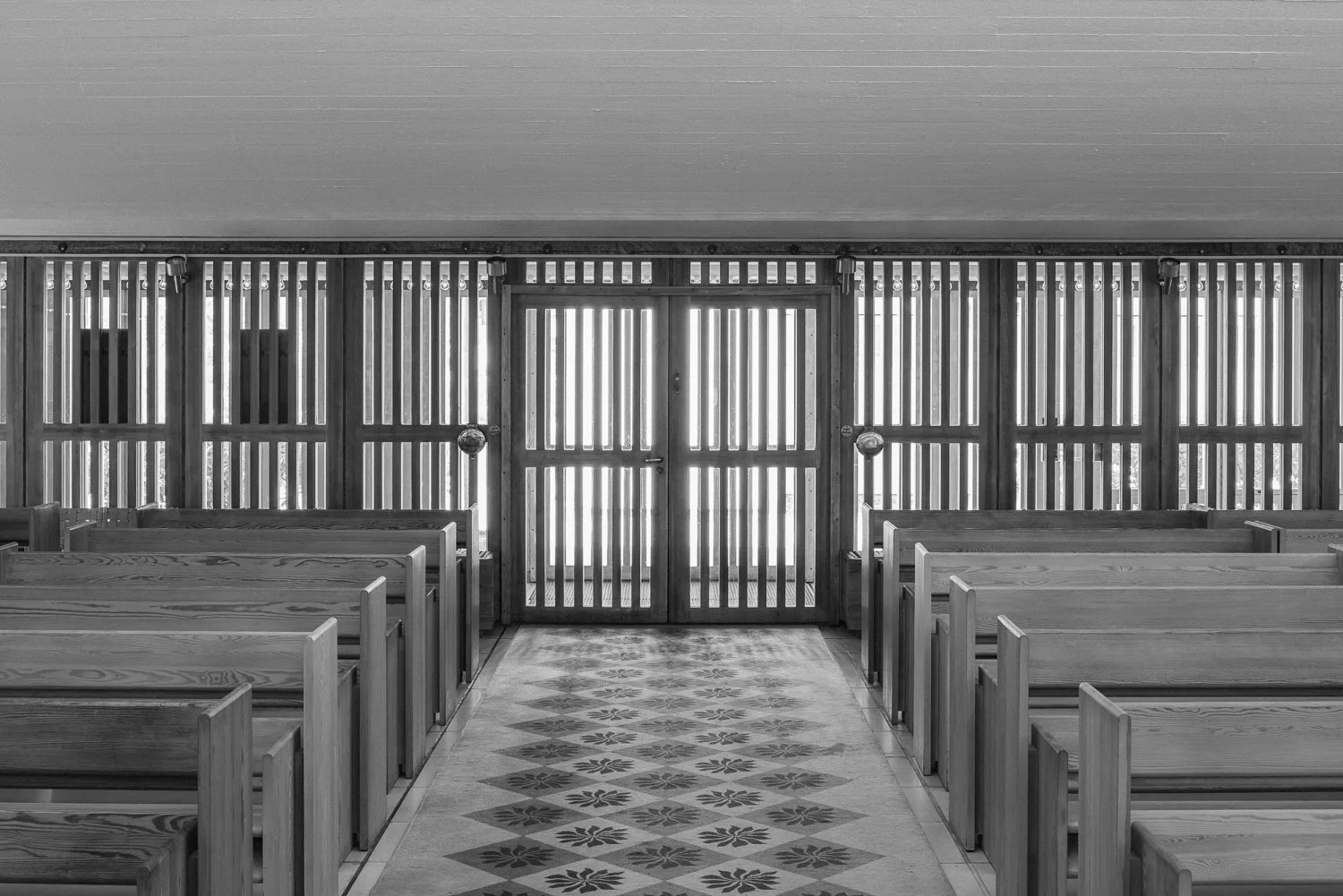 Black and white image of interior of a wooden chapel with a central door flanked by wooden slat walls, wooden benches arranged on both sides, and a patterned tile floor.
