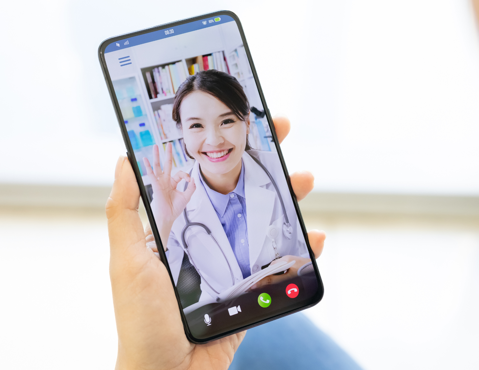 Person holding smartphone displaying video call with smiling doctor, wearing white coat and stethoscope, making OK gesture.