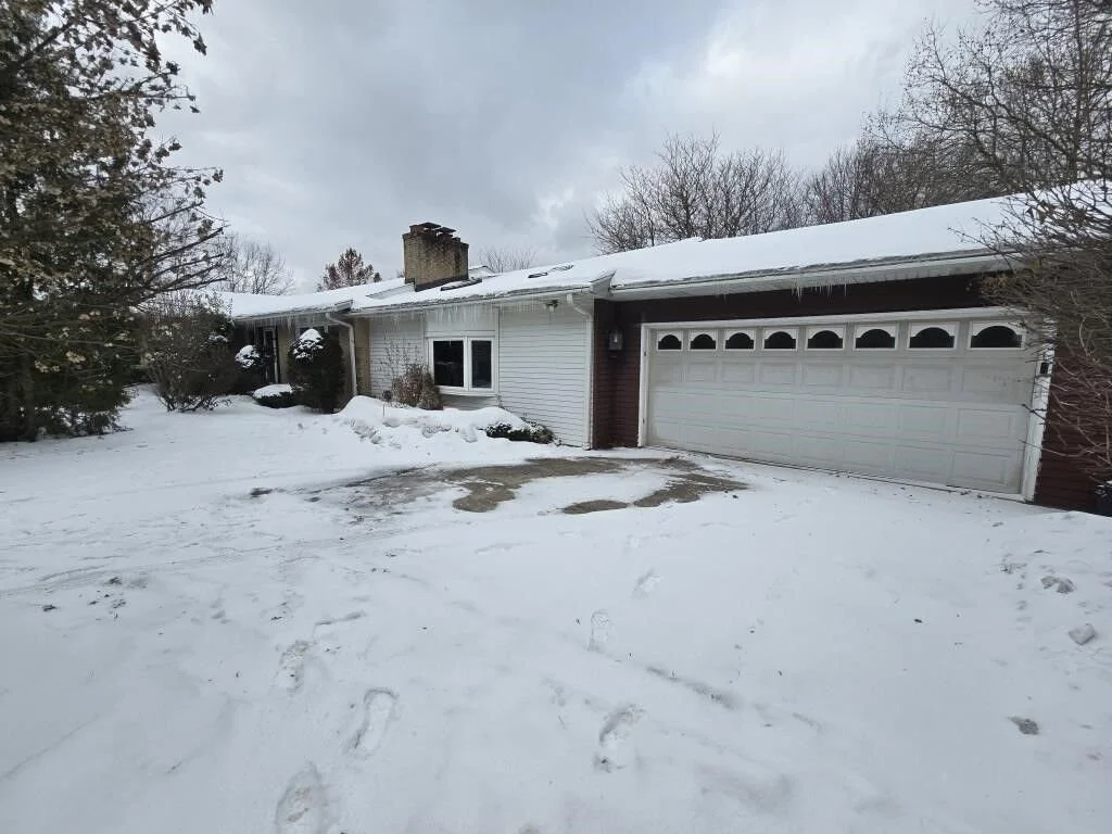 A house with white siding and a two-car garage, surrounded by snow with footprints and tire tracks, leafless trees, and an overcast sky.