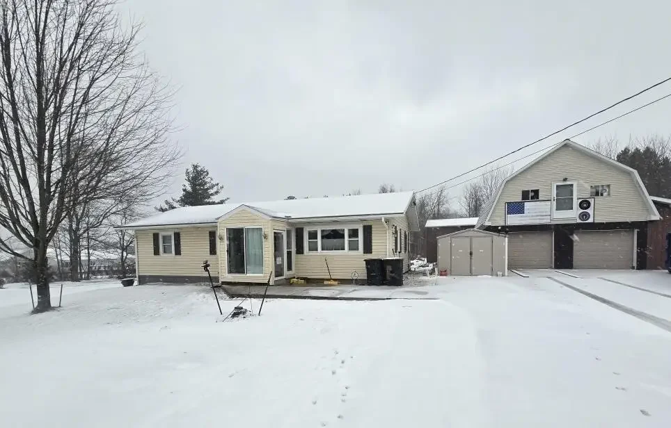 Snow-covered backyard with a one-story house on the left and a two-story garage and house on the right, with snow and overcast sky