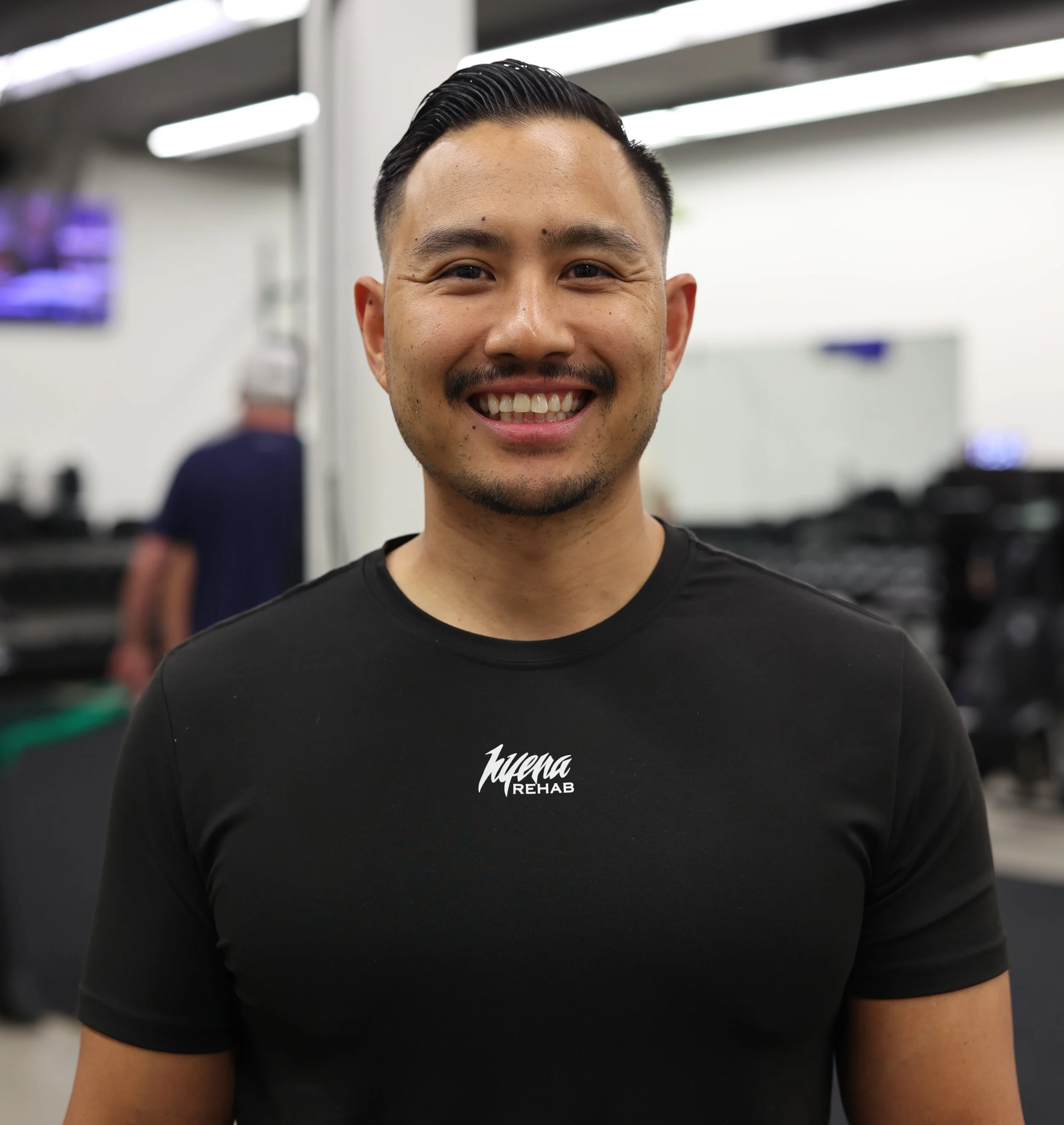 A smiling man with slicked-back black hair and a mustache, wearing a black T-shirt with "Huyena REHAB" written on it, in a gym or rehabilitation center. Background shows blurred people and equipment.