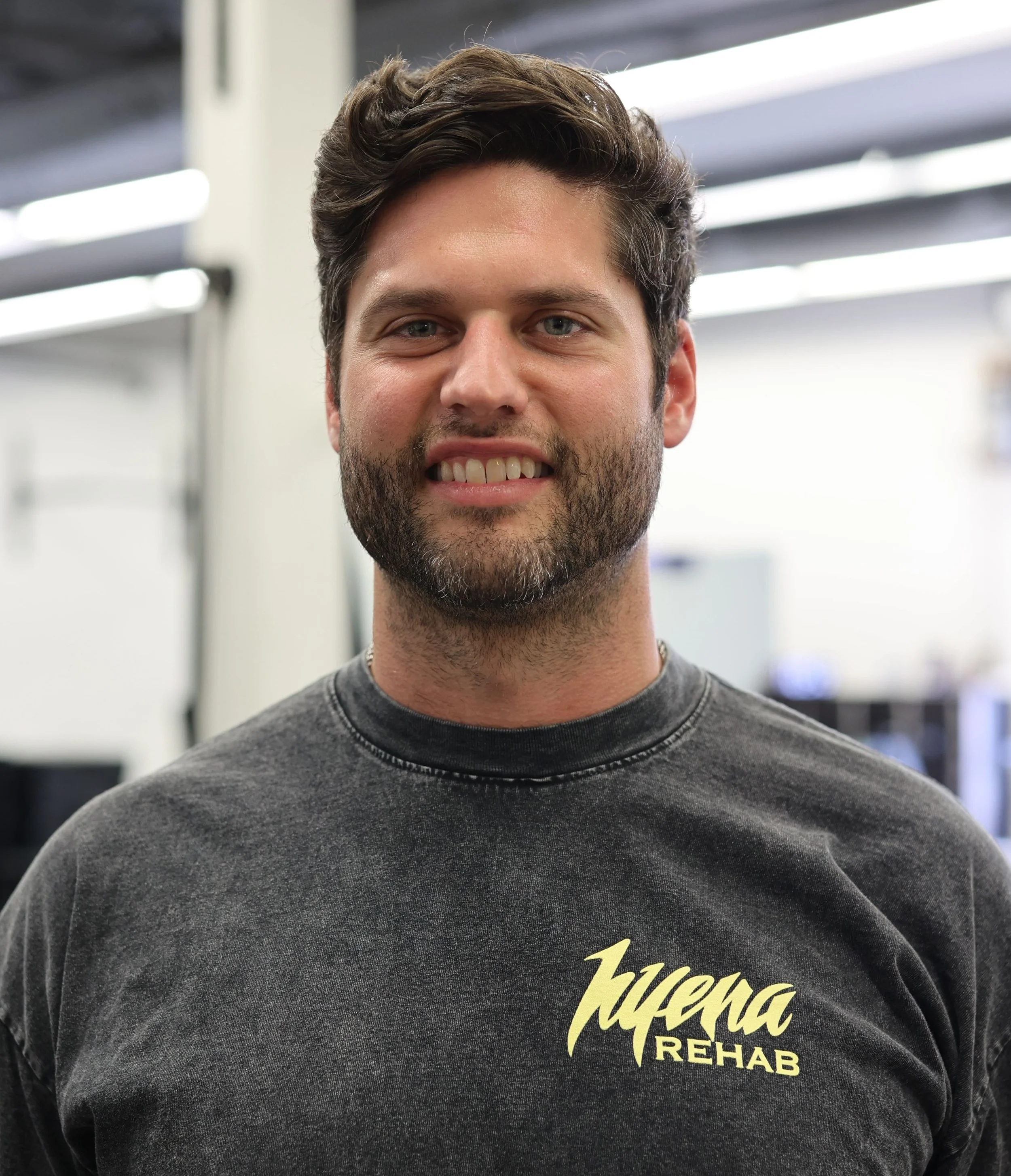 A man with dark hair and a beard wearing a black T-shirt that says Juvena REHAB in yellow letters, smiling inside a gym or workout facility.