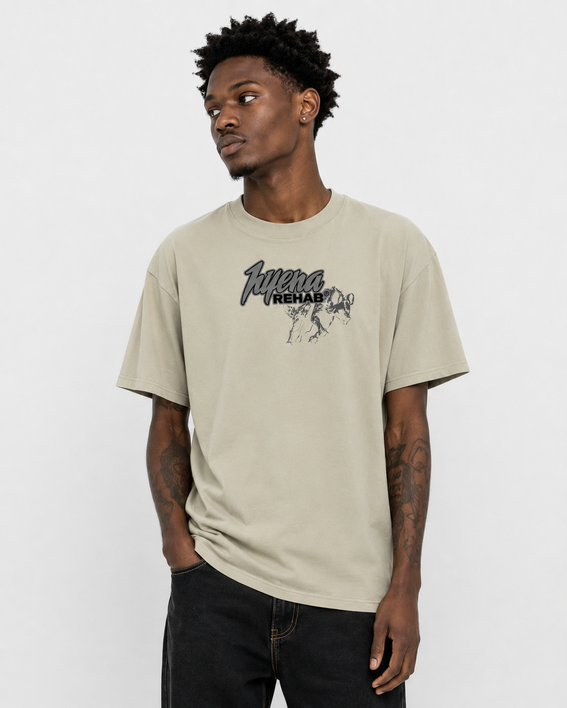 A young man with dark skin and curly black hair is wearing a beige T-shirt with the words 'Aura Rehab' and a graphic of a dog on it. He has tattoos on his arms and is standing against a plain white background.