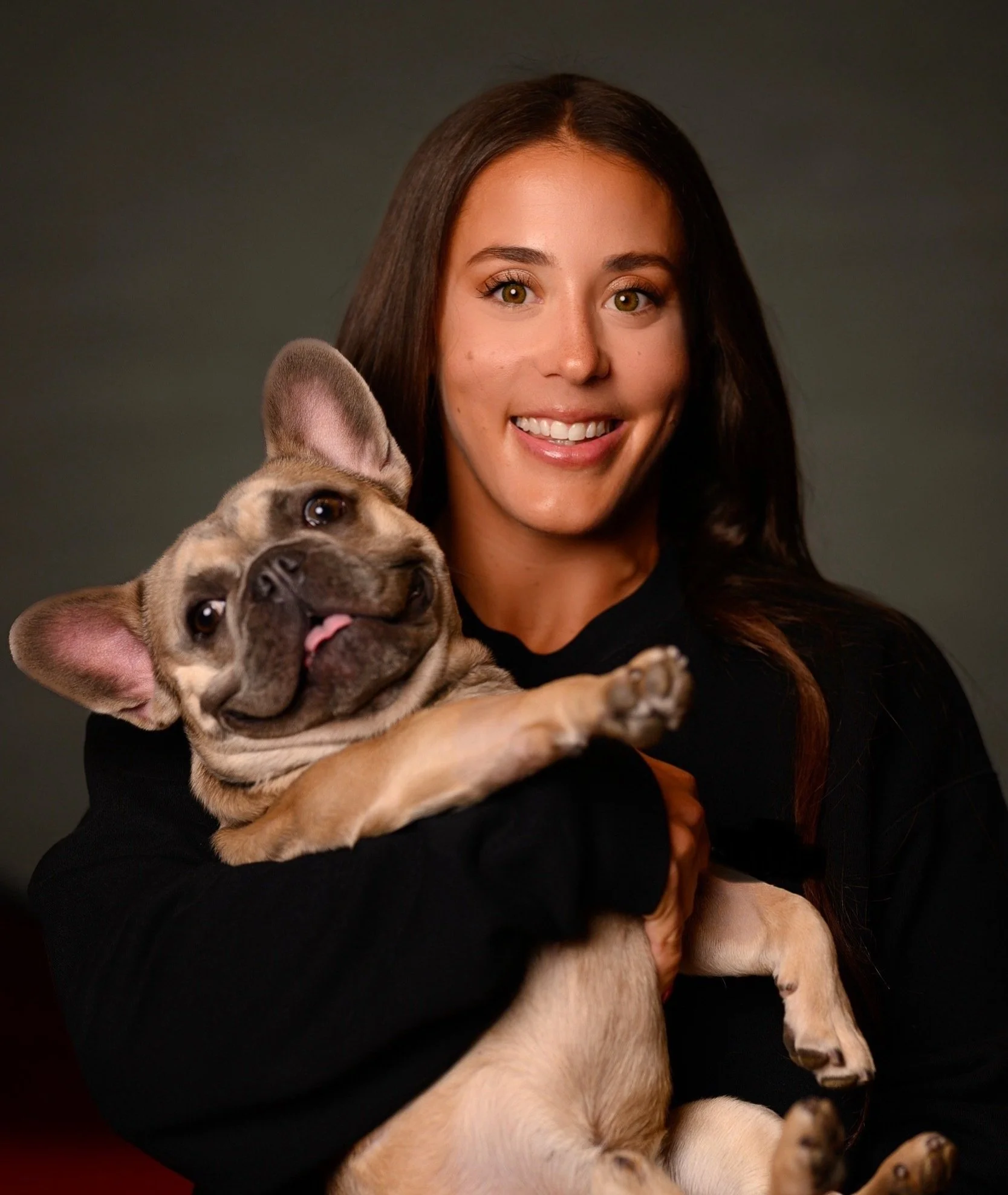A woman with long dark hair smiling while holding a French Bulldog puppy.