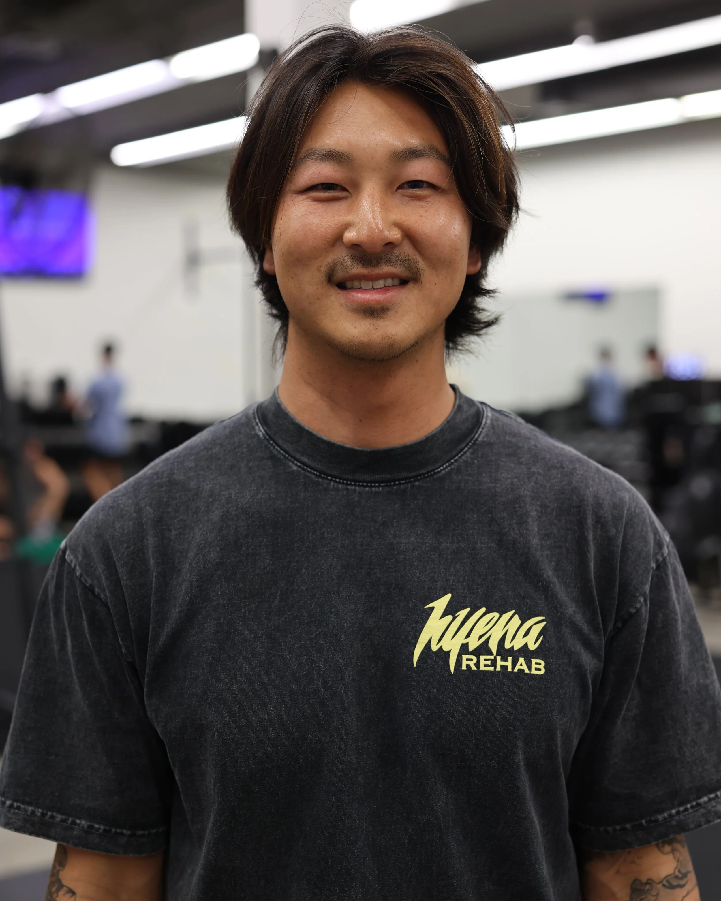 A man with medium-length dark hair smiling at the camera, wearing a black T-shirt with yellow text that reads "Lucera REHAB" inside a gym or fitness center with people working out in the background.