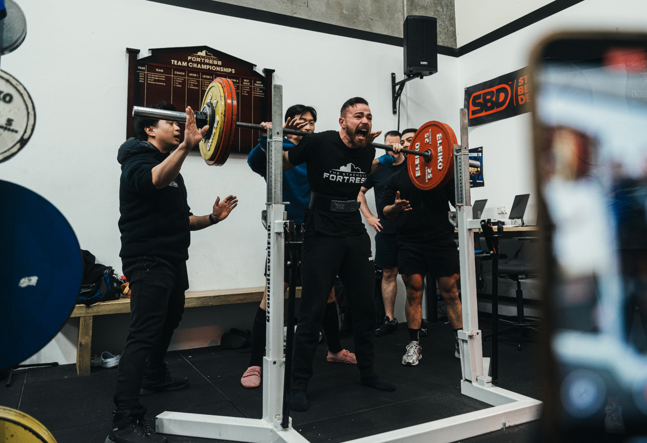 A male lifter squats a PB. He is surrounded by his friends who are spotting him