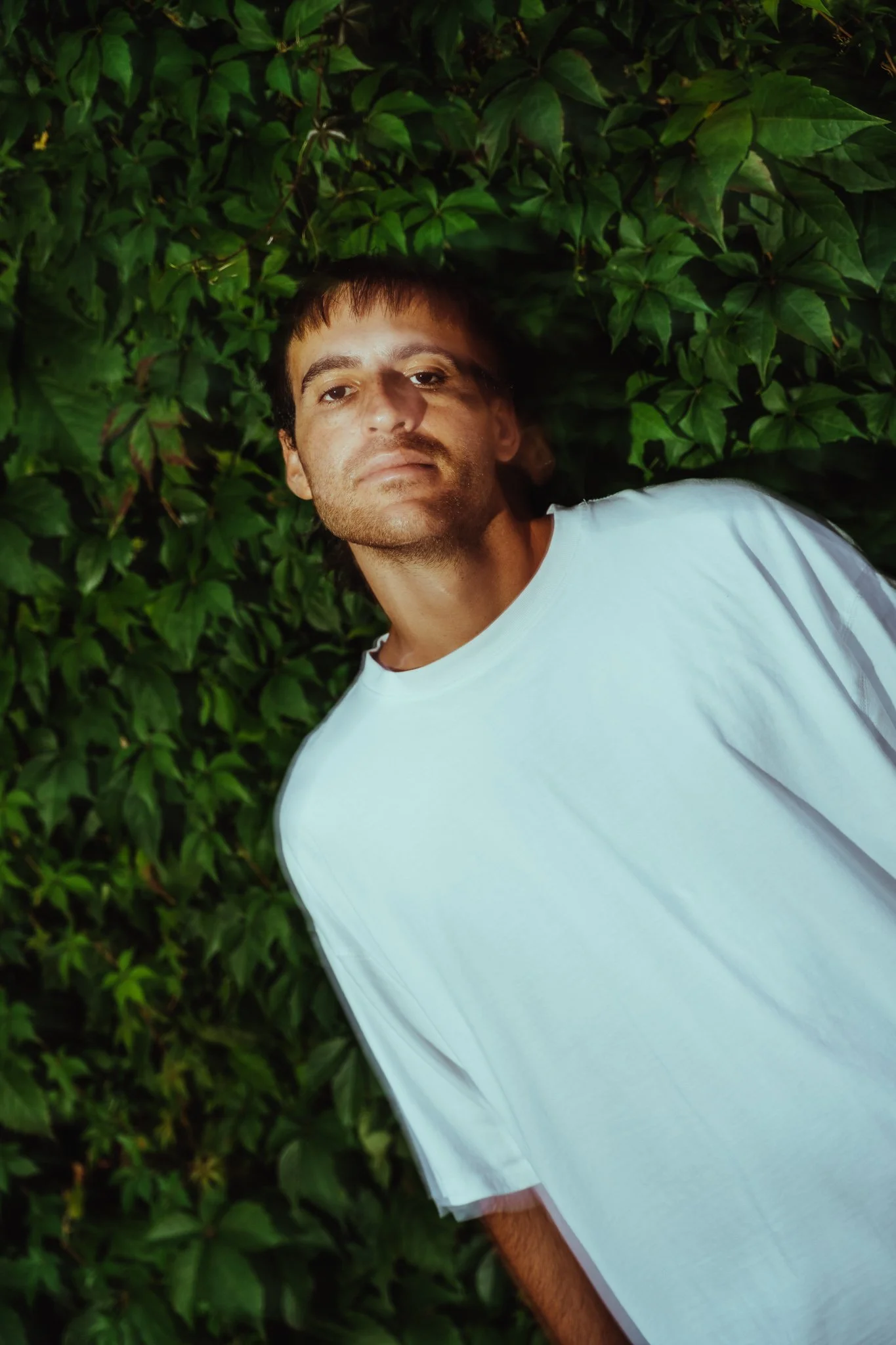 A man with short dark hair and light skin lying on the ground surrounded by green leaves, wearing a white T-shirt.