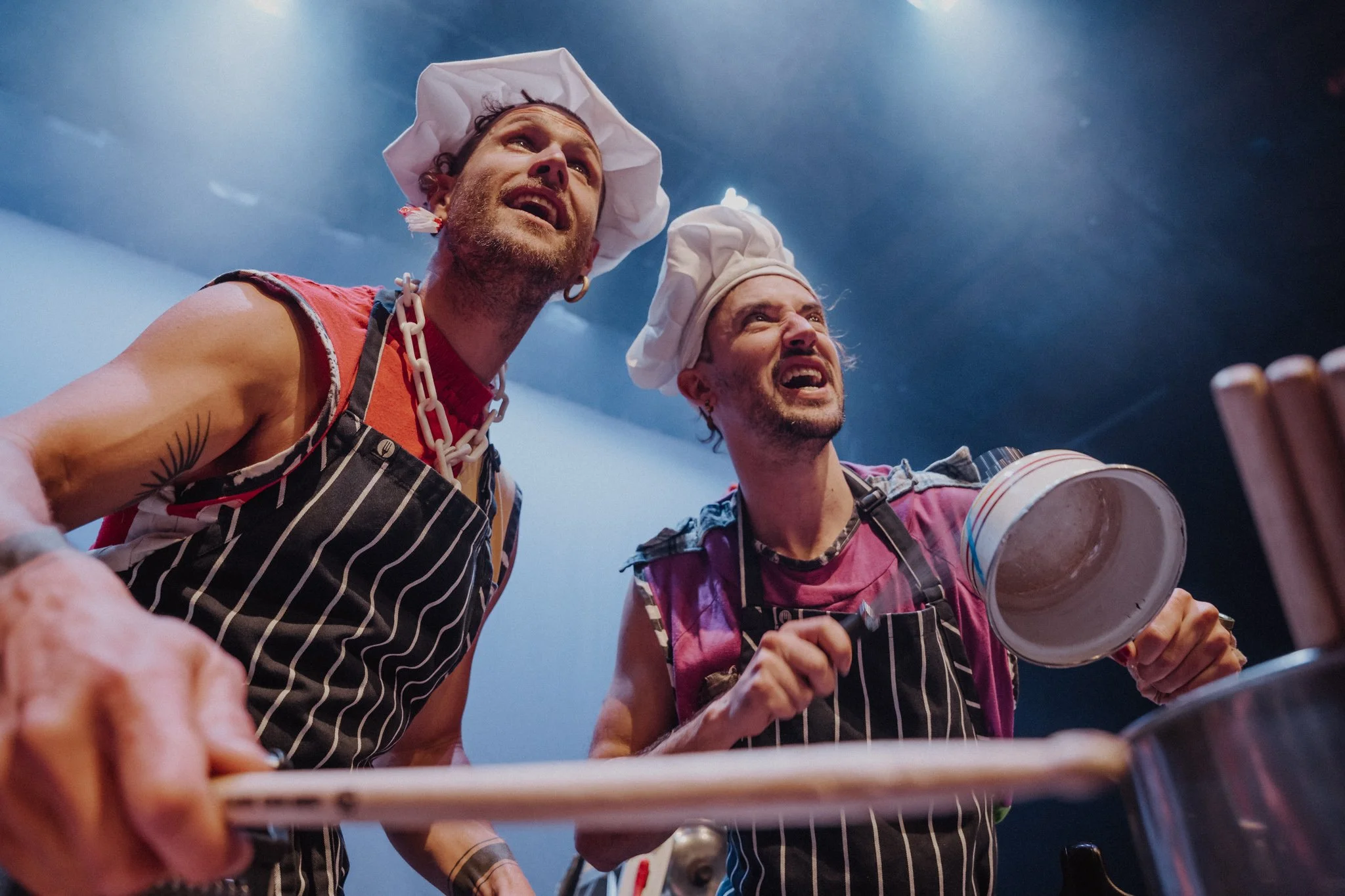 Two men dressed as chefs wearing chef hats and striped aprons look up with expressions of concern or concentration while cooking in a kitchen.