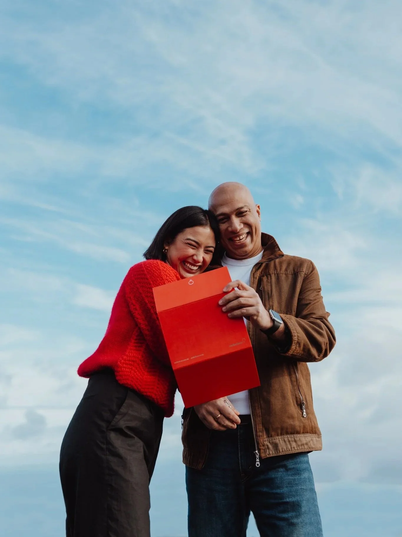 A smiling couple looking at a red box together outdoors against a blue sky.