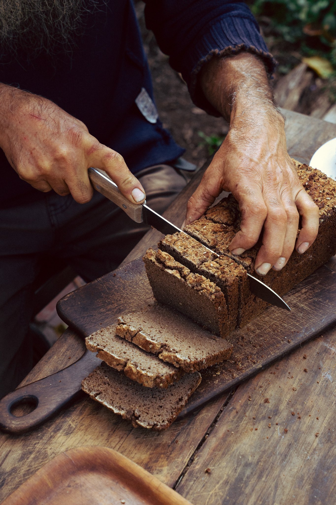 A person is slicing a loaf of homemade bread on a wooden cutting board outdoors.