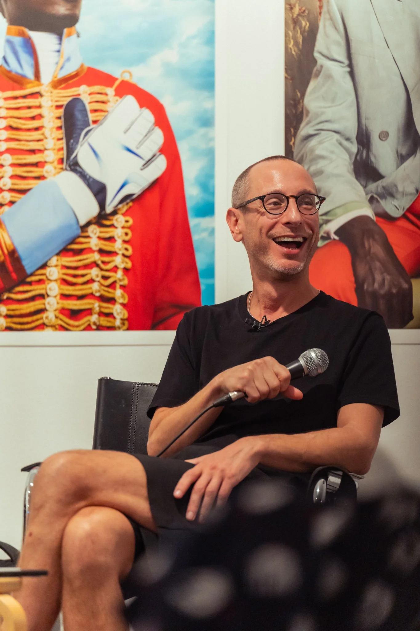 A man sitting in a chair, holding a microphone, smiling and wearing glasses with a black t-shirt and shorts, with large, colorful portrait photos in the background.