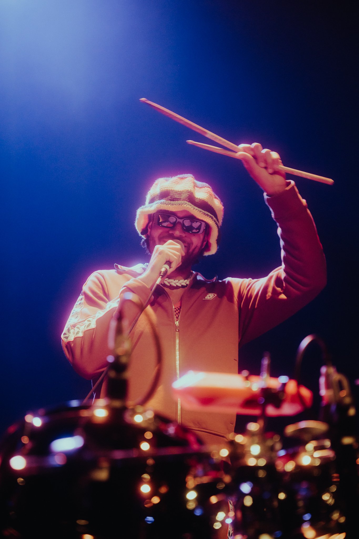 A performer on stage holding drumsticks in the air, wearing sunglasses, a beanie, and an orange jacket, with drums and equipment in front of him, under colorful stage lighting.
