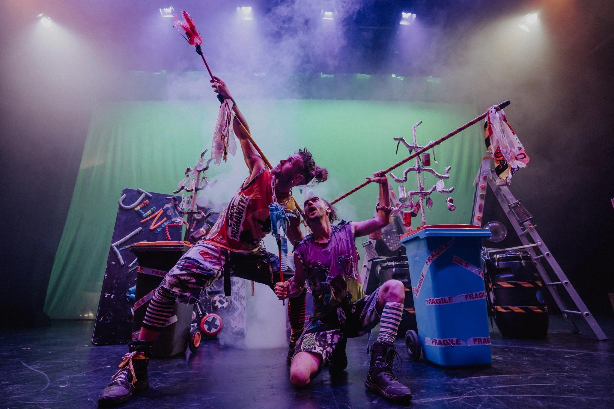 Two performers with colorful, chaotic costumes on a stage filled with assorted props like a trash bin, ladder, and toys. They hold long sticks and appear to be acting out a dramatic, theatrical scene with a smoky, vibrant background.