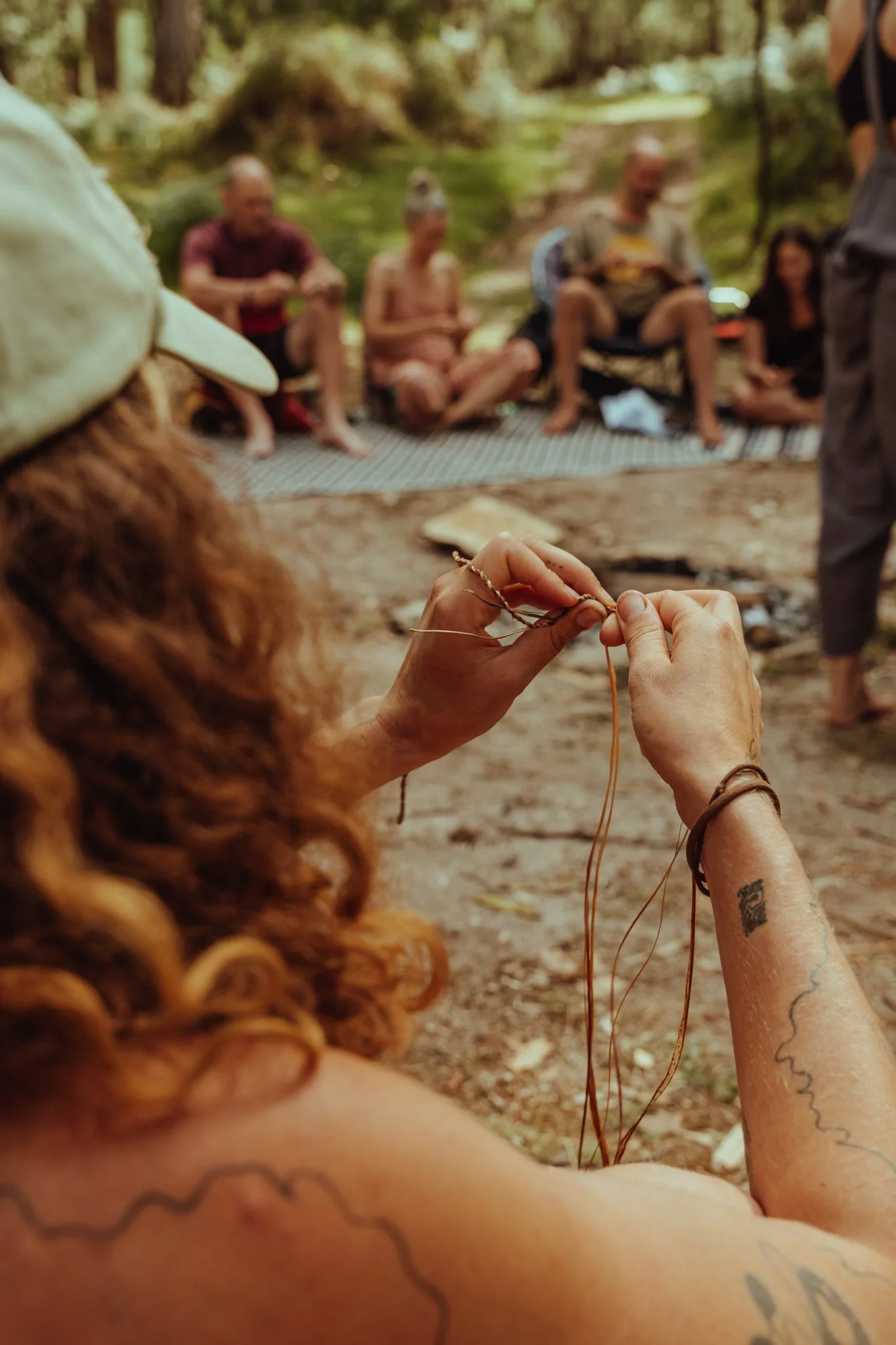 Close-up of a woman with curly red hair and tattoos on her arms, sitting outdoors, making a bracelet with brown string. In the background, a group of five people sit on a blanket, with trees and greenery surrounding them, indicating a nature setting.