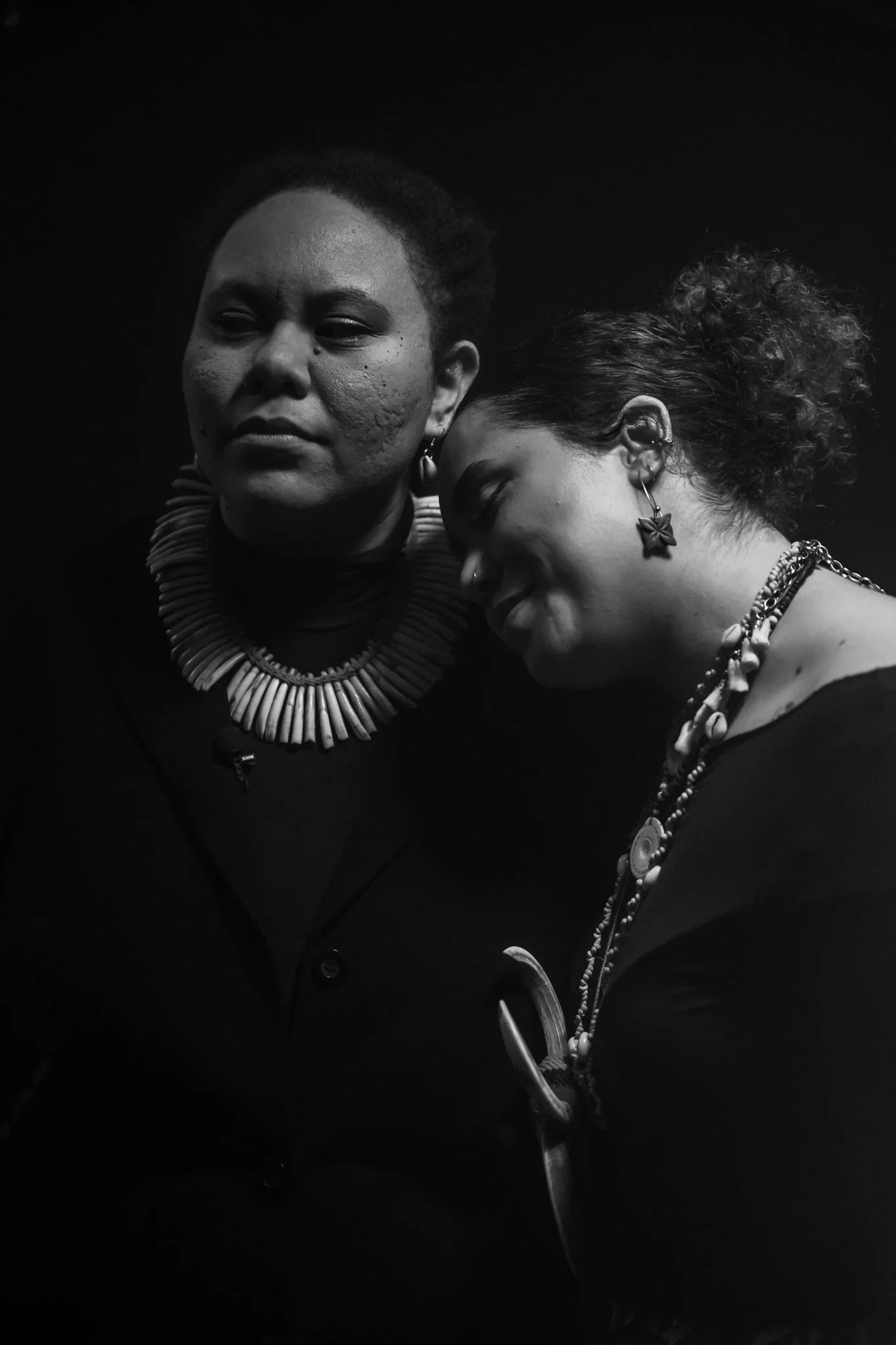 Two women with dark skin, jewelry, and earrings are depicted in a black-and-white photo, with one woman resting her head on the other's shoulder.