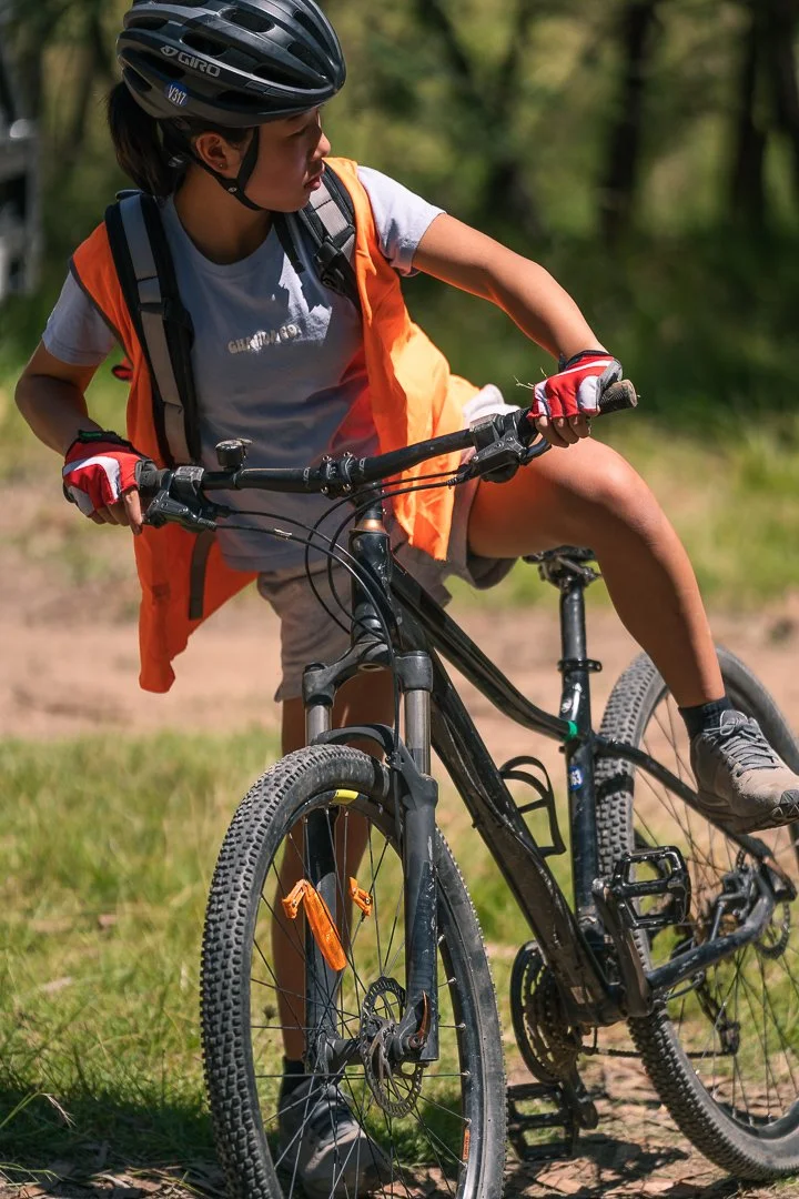 Young girl wearing a helmet and gloves riding a mountain bike outdoors on a dirt trail with green trees in the background.