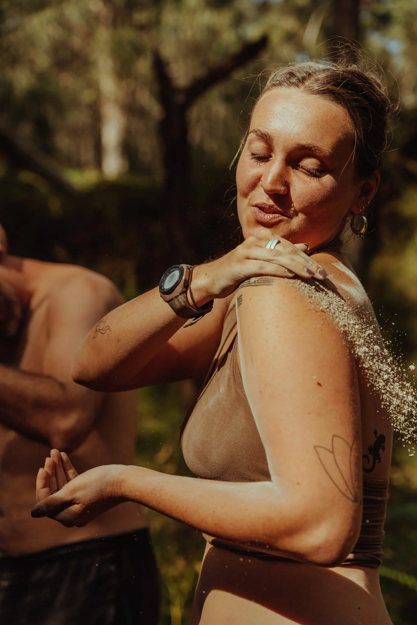 A young woman with tattoos and piercings, wearing a sleeveless top, standing outdoors in a wooded area. She appears to be enjoying a moment, possibly after a workout or a dance, with her eyes closed and a relaxed expression. Sand is visible on her sh