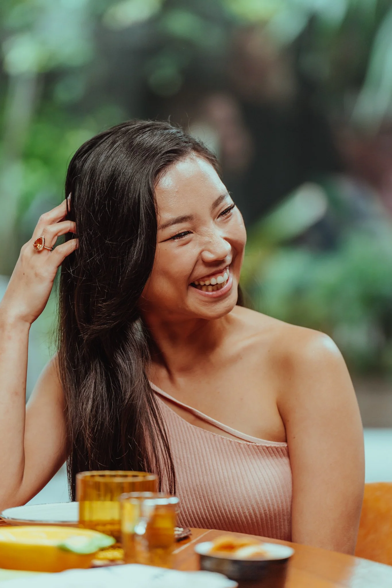 A woman smiling and laughing while sitting at a table with dishes and glasses, with a blurred green outdoor background.