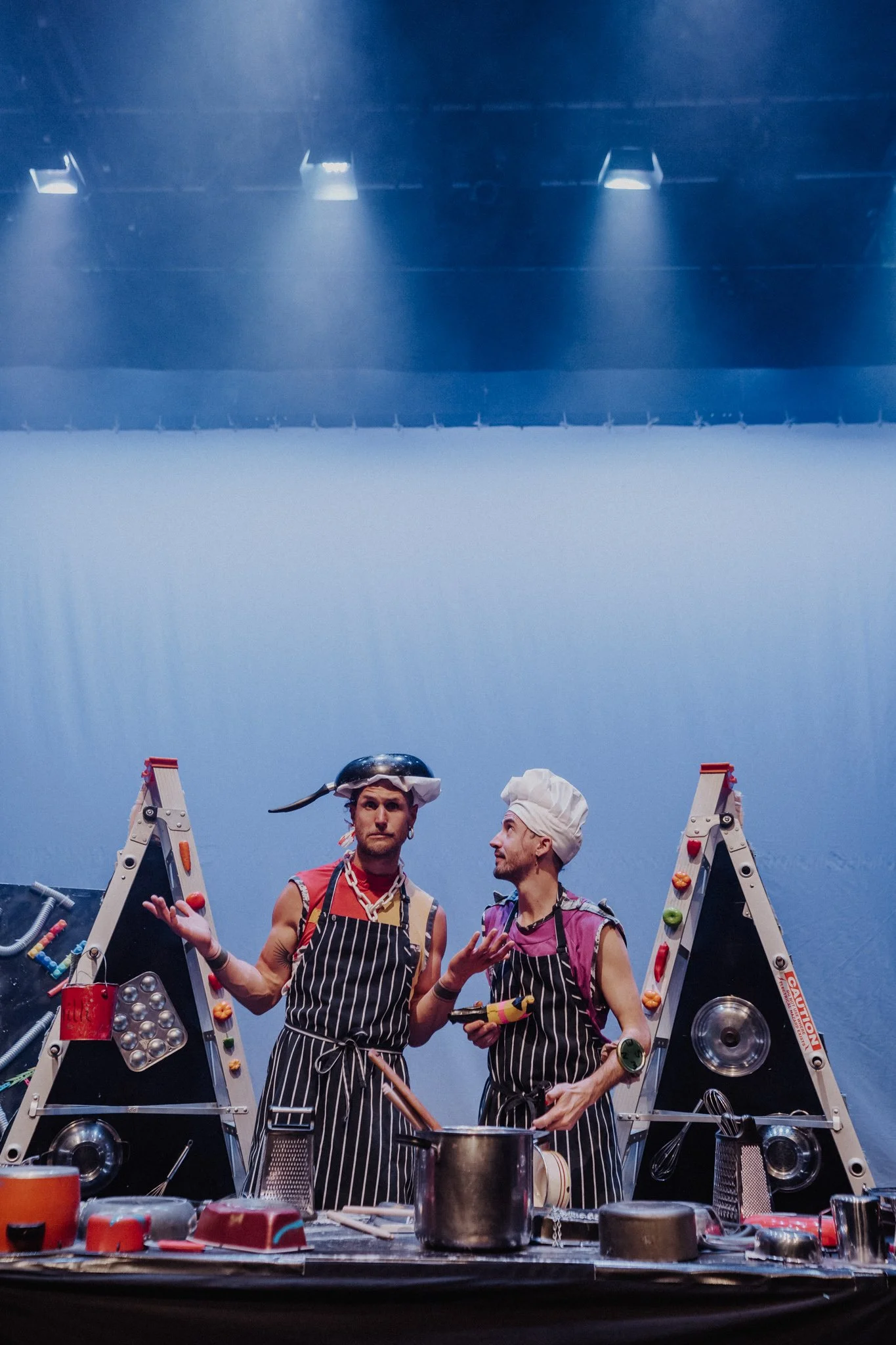 Two men dressed as chefs standing behind a cluttered table with kitchen tools and ladders, on a stage with blue lighting.