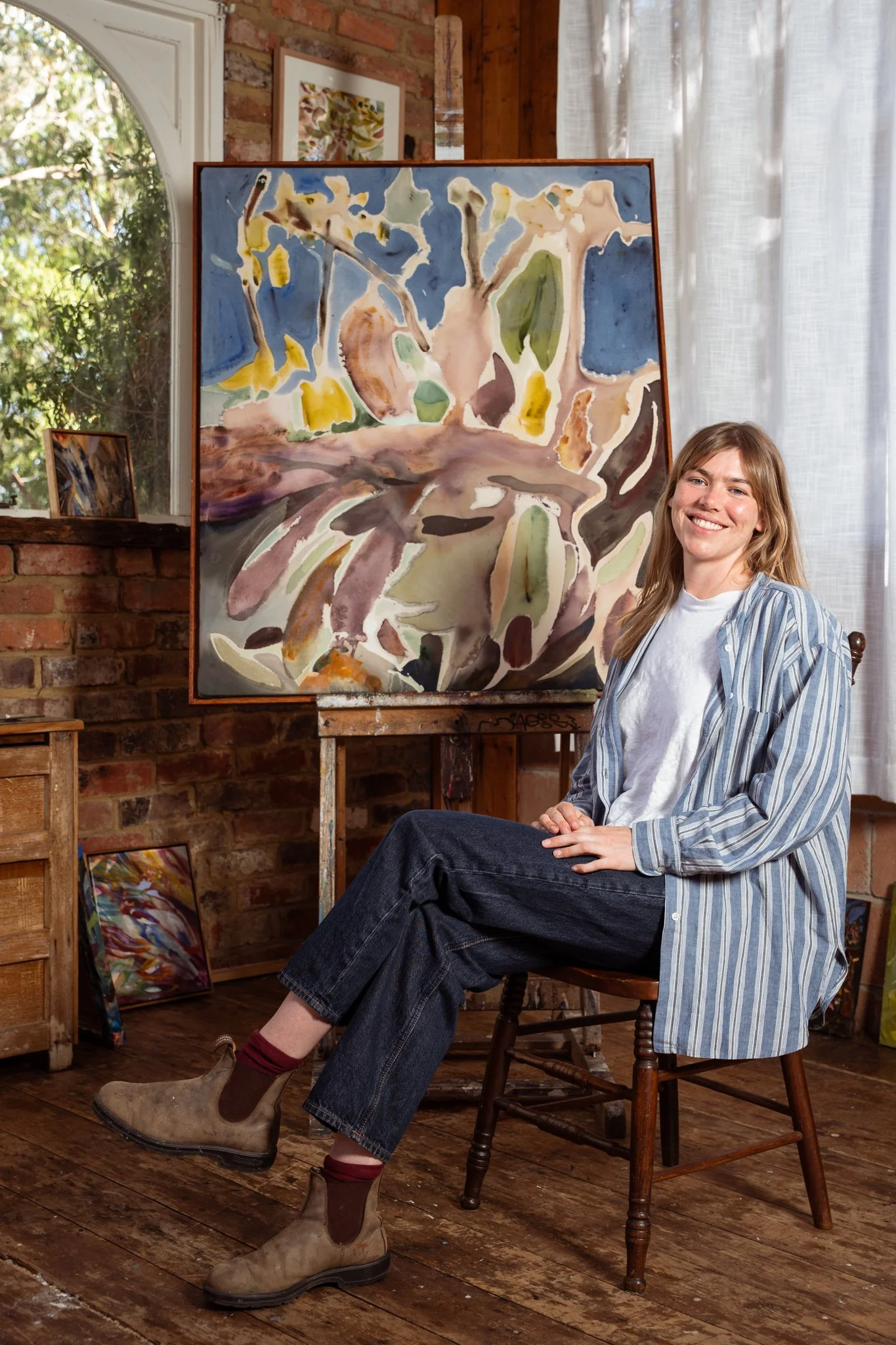 Young woman sitting on a wooden chair in an art studio, smiling, wearing a striped shirt, white t-shirt, jeans, and boots. Behind her is a large abstract watercolor painting on an easel, with smaller paintings on the floor and wall, and a brick wall 