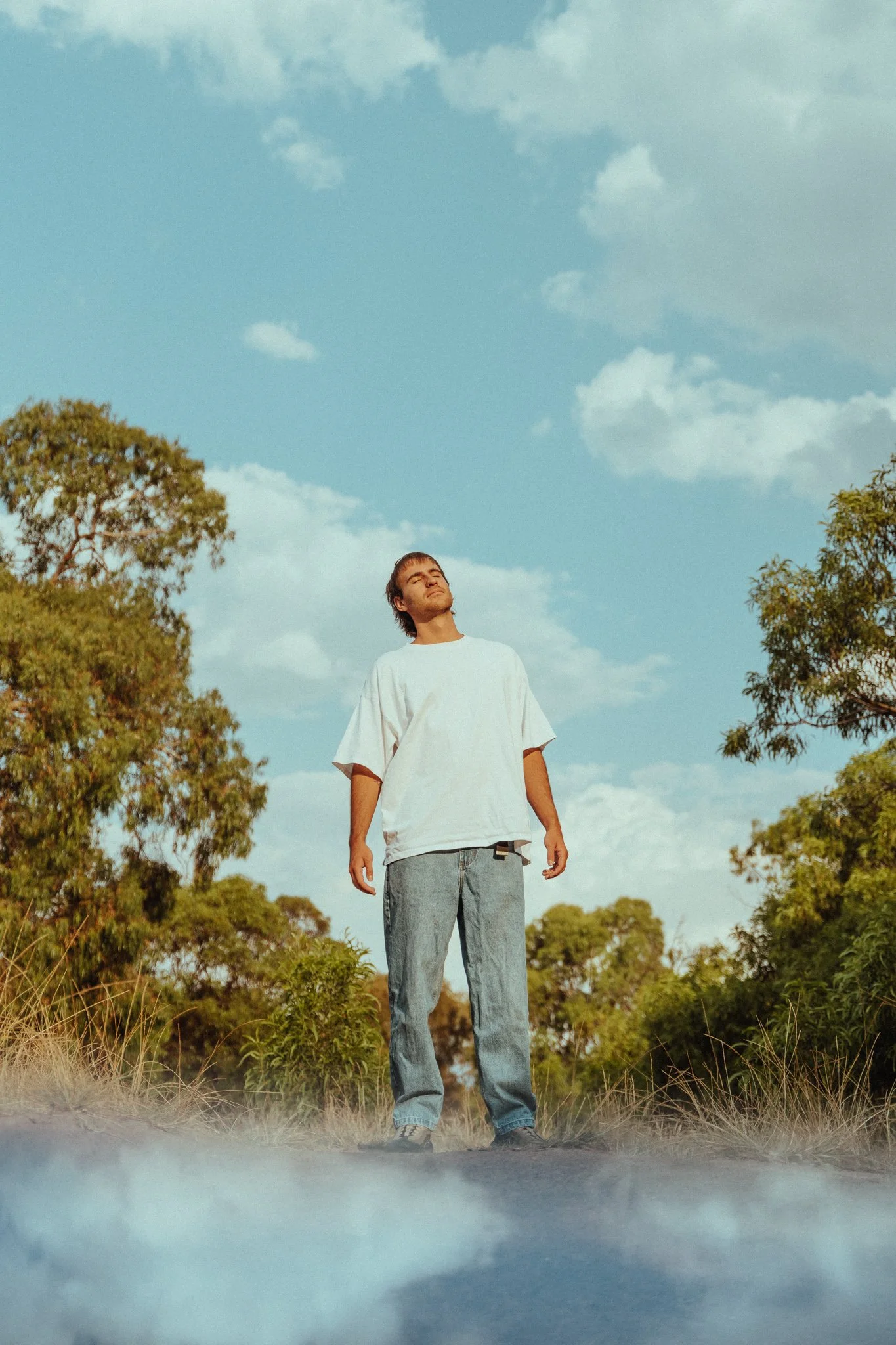 Young man standing outdoors with eyes closed, facing upward, surrounded by trees and a bright sky.