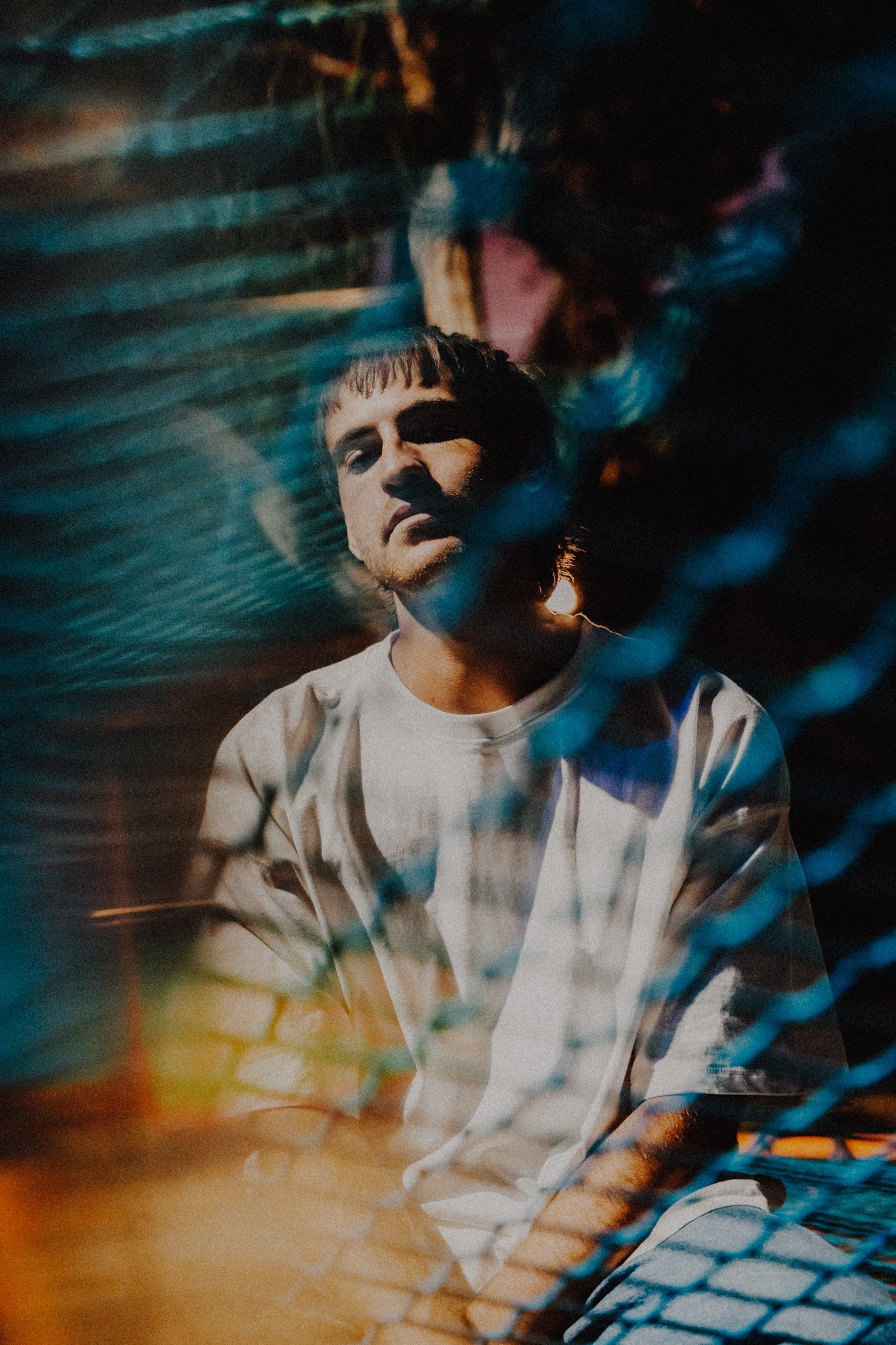 A young man with short brown hair and a beard is sitting behind a chain-link fence, with colorful lights creating a moody atmosphere at night.