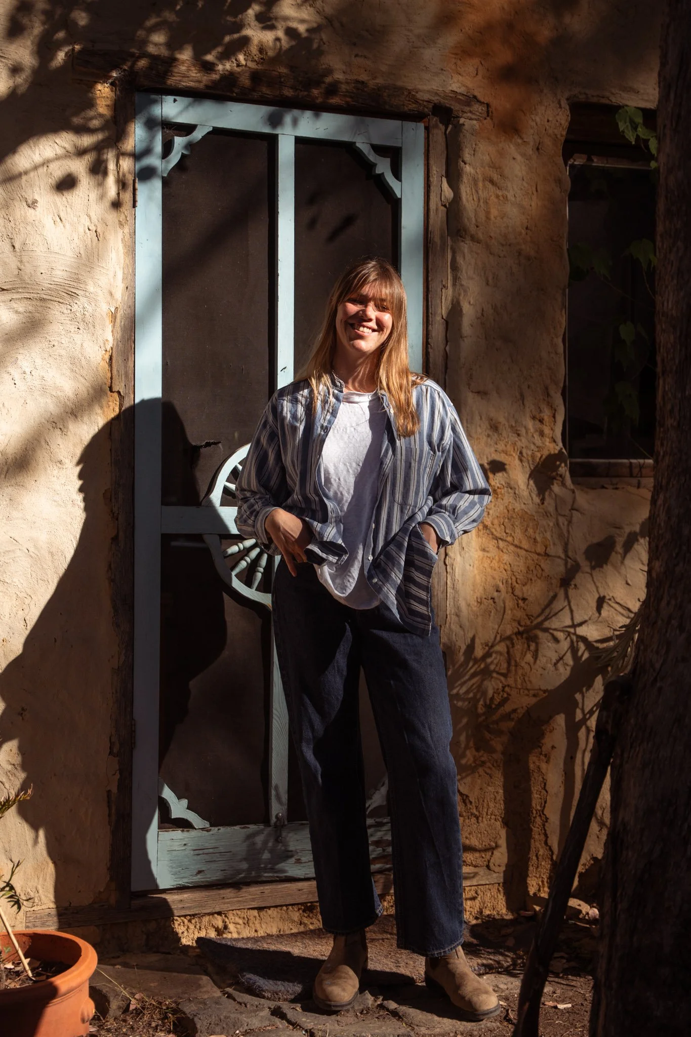 A woman with blonde hair, wearing a striped shirt over a white t-shirt, dark jeans, and boots, standing outdoors in front of a rustic door and smiling.