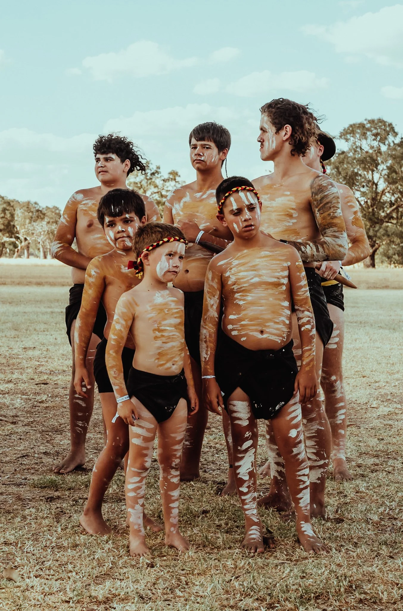 Group of children and young adults with body paint, wearing black shorts, standing outdoors on a grassy field under a blue sky with clouds.