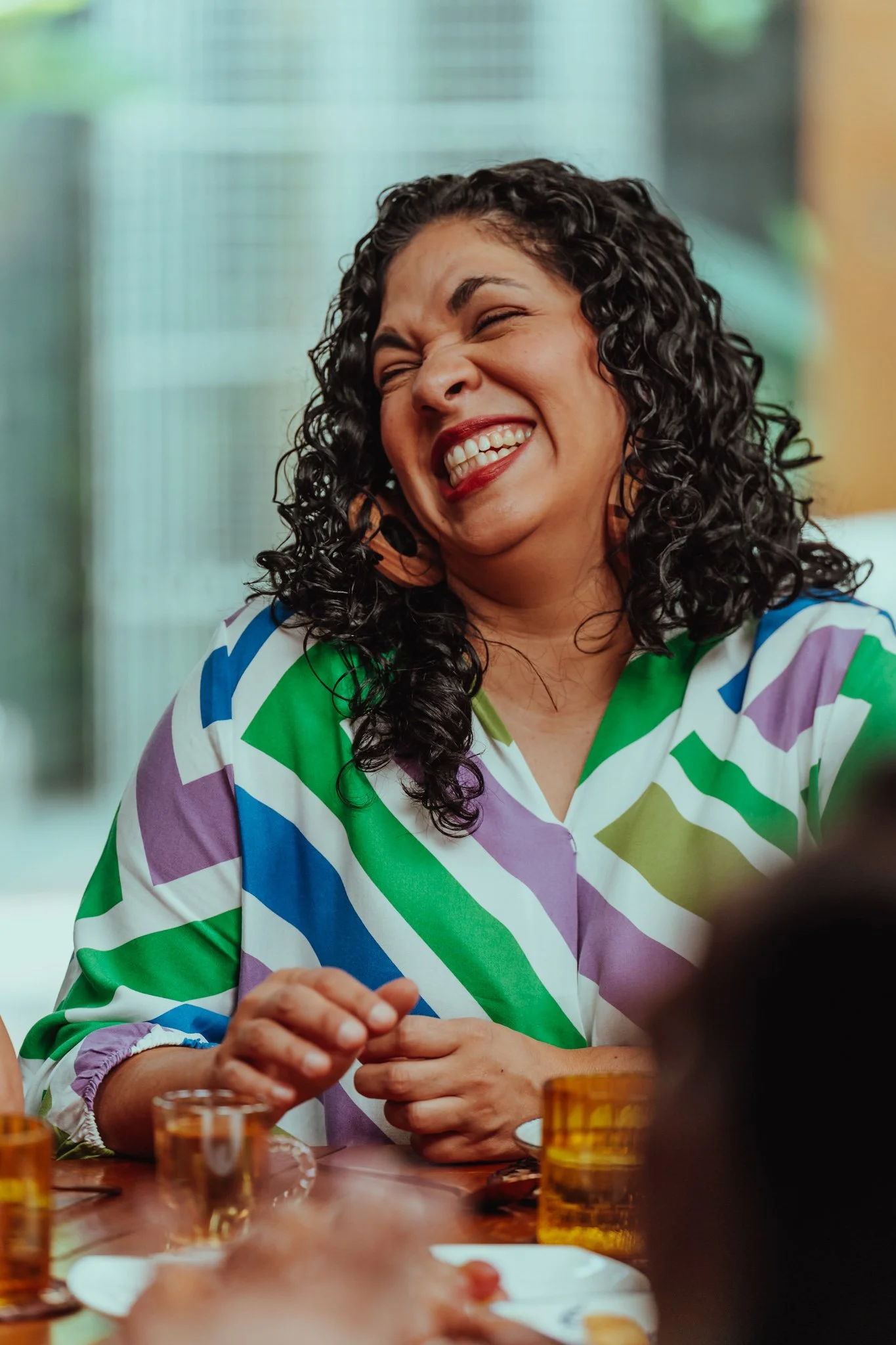 A woman with curly dark hair is smiling and laughing at a table with food and drinks.