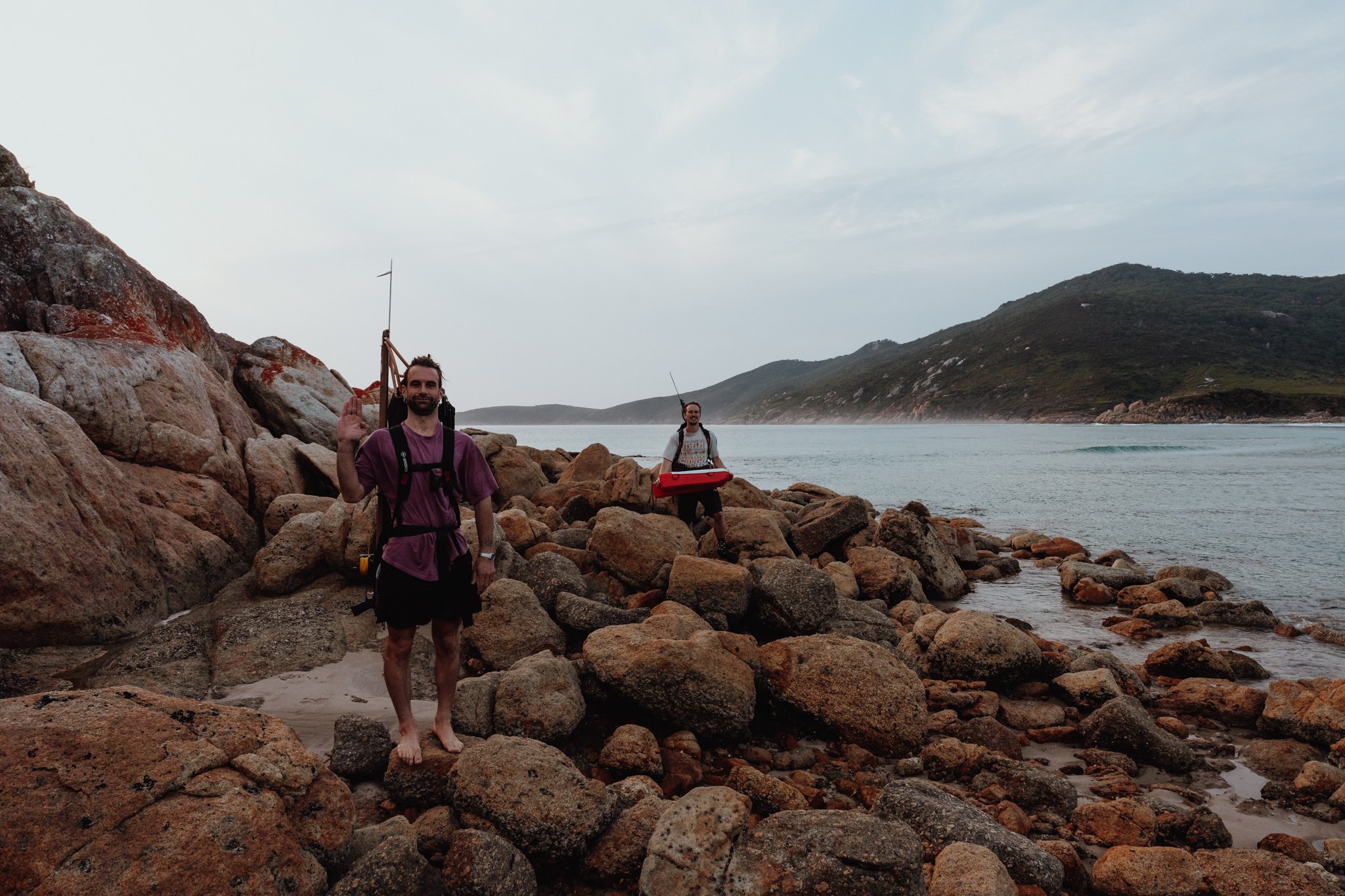 Two men with backpacks and fishing gear on a rocky shoreline near the ocean, with one waving and the other holding a red tackle box, and hills in the background.
