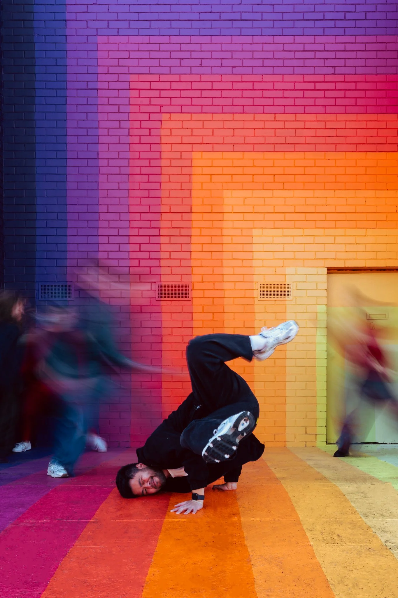 A man breakdancing on a colorful rainbow-painted sidewalk with a vibrant rainbow-colored brick wall in the background. Several blurred people are walking past him.
