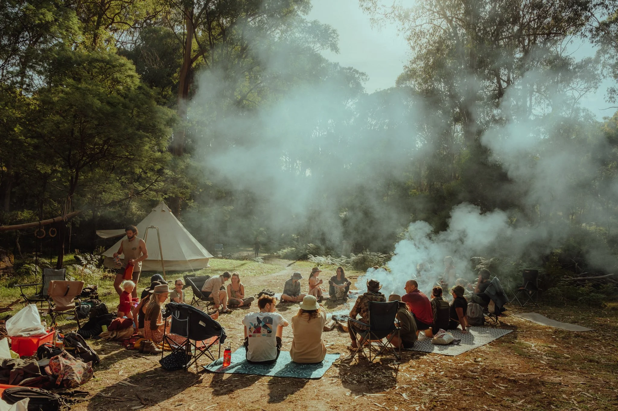 Group of people gathered around a campfire in a forested area, with smoke rising, tents in the background, and trees surrounding the scene.