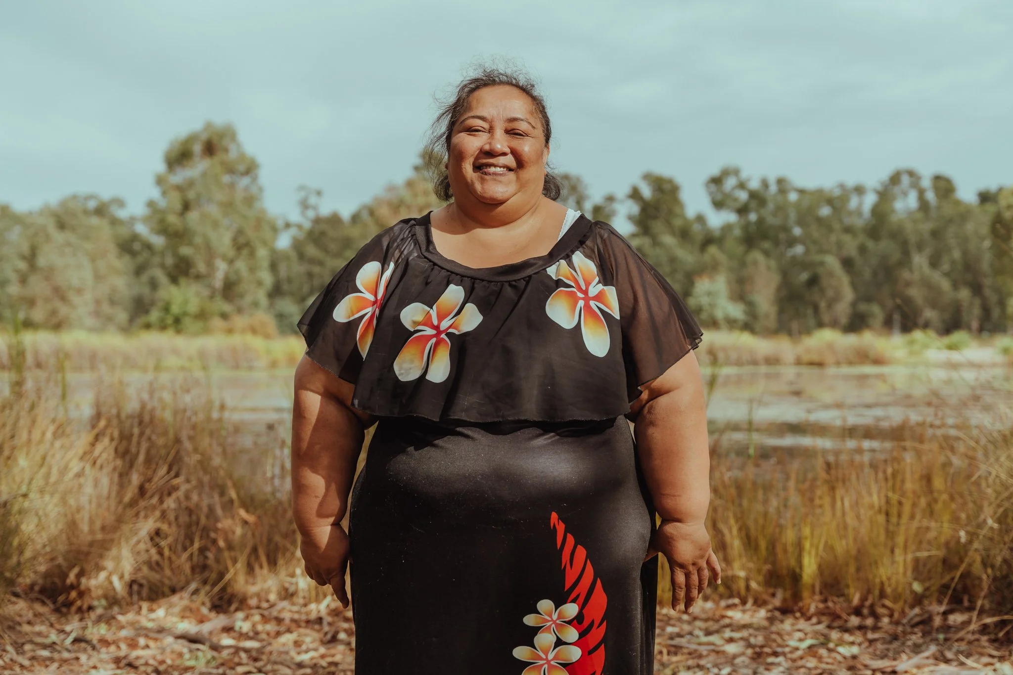 A smiling woman standing outdoors in front of a lake and trees, wearing a black dress with floral designs and a black shawl with flower print.